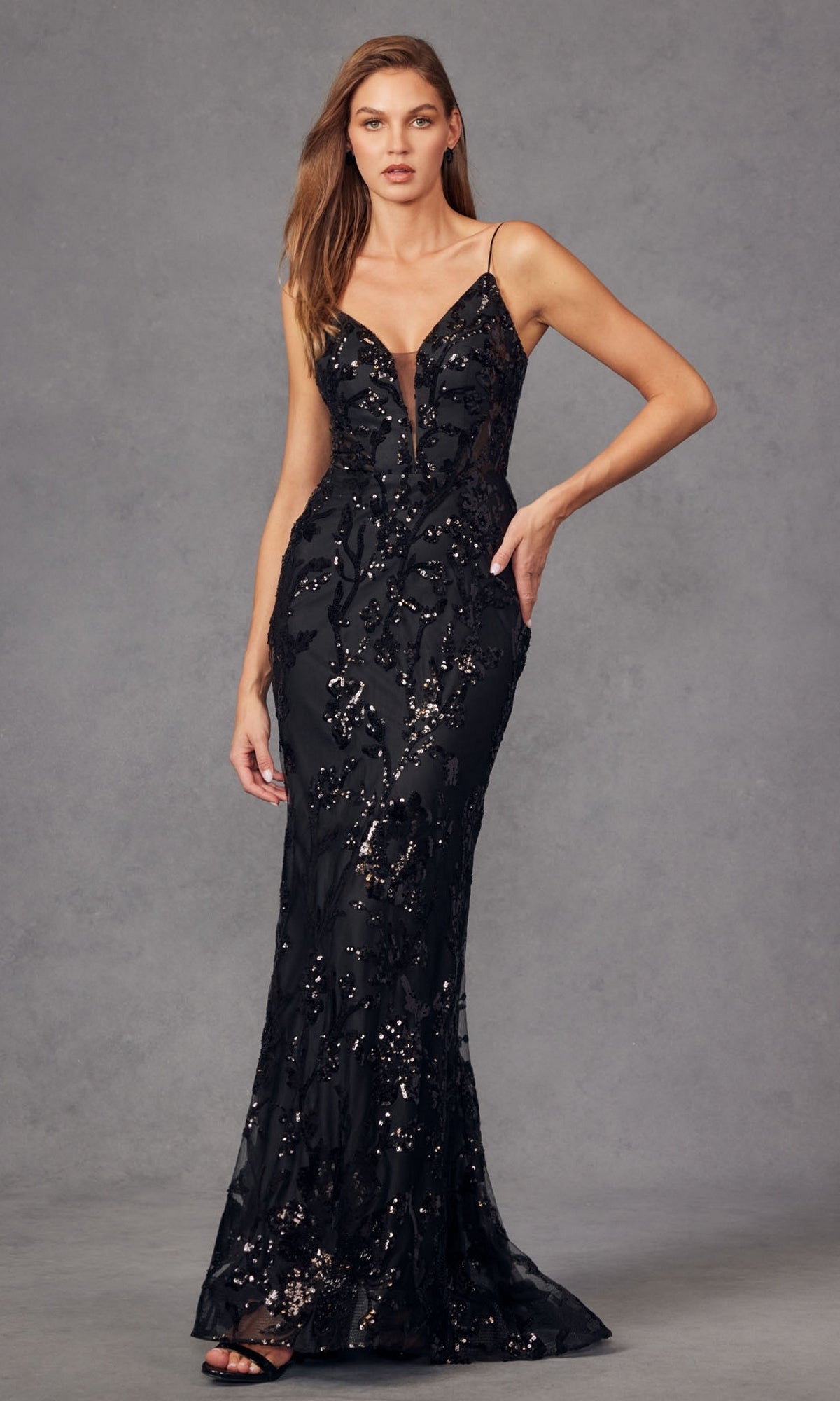 A woman poses against a gray backdrop wearing the Strappy-Back Long Sequin-Print Prom Dress 2411, a floor-length black dress with sequins, thin straps, an open back, and a deep V neckline.
