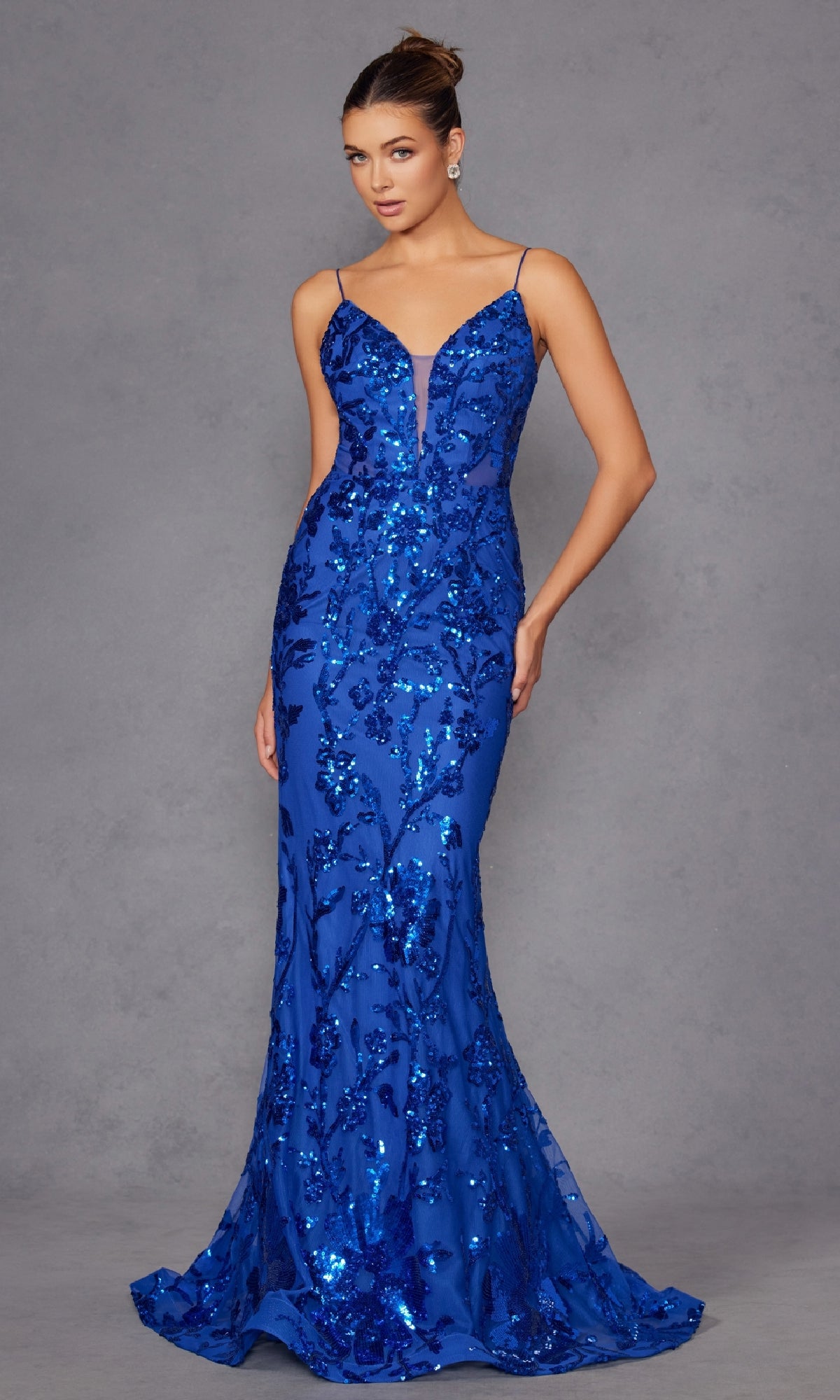 A woman poses against a plain gray background, wearing the Strappy-Back Long Sequin-Print Prom Dress 2411—a fitted, floor-length royal blue gown adorned with sequins, featuring thin straps and a deep V neckline.