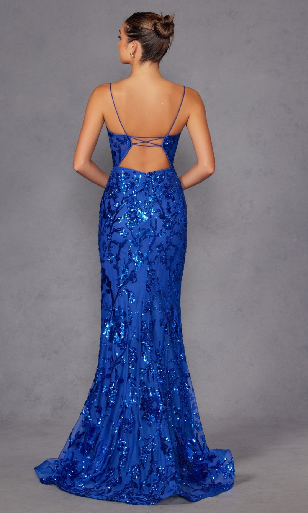 A woman faces away, wearing the Strappy-Back Long Sequin-Print Prom Dress 2411—a floor-length, fitted royal blue gown with an open strappy back—set against a plain gray backdrop; perfect for formal evenings.