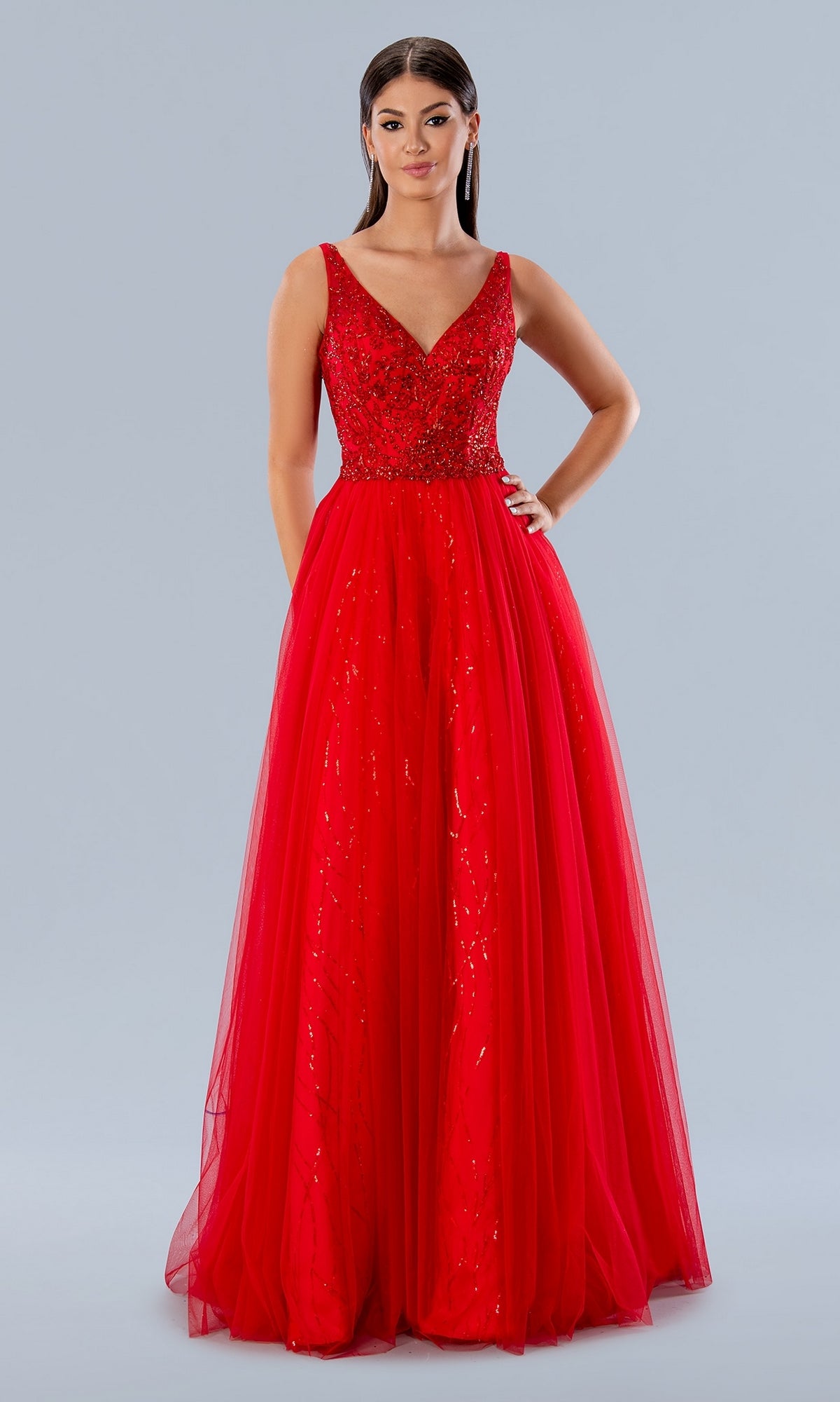 A woman wears the Stella Couture 24150 V-Neck Long Tulle Prom Dress, a sleeveless red gown with a fitted, embellished bodice and full tulle skirt, standing against a plain light gray background.