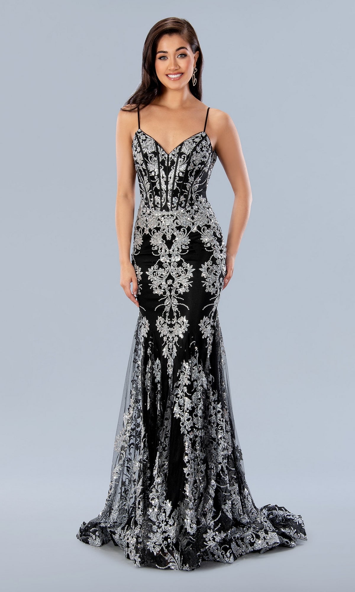 A woman wearing the Long Prom Dress 24159 by Stella Couture—a black mermaid gown with metallic embroidery, thin straps, and a fitted bodice—stands against a plain gray background.