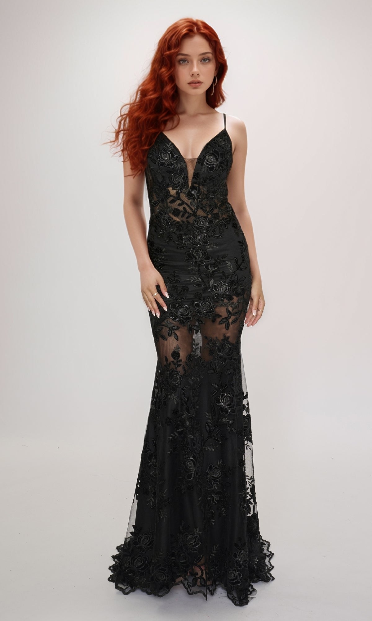 Woman with long red hair wearing the black, floor-length lace Nina Canacci 2429 formal dress with sheer panels, standing against a plain light background.