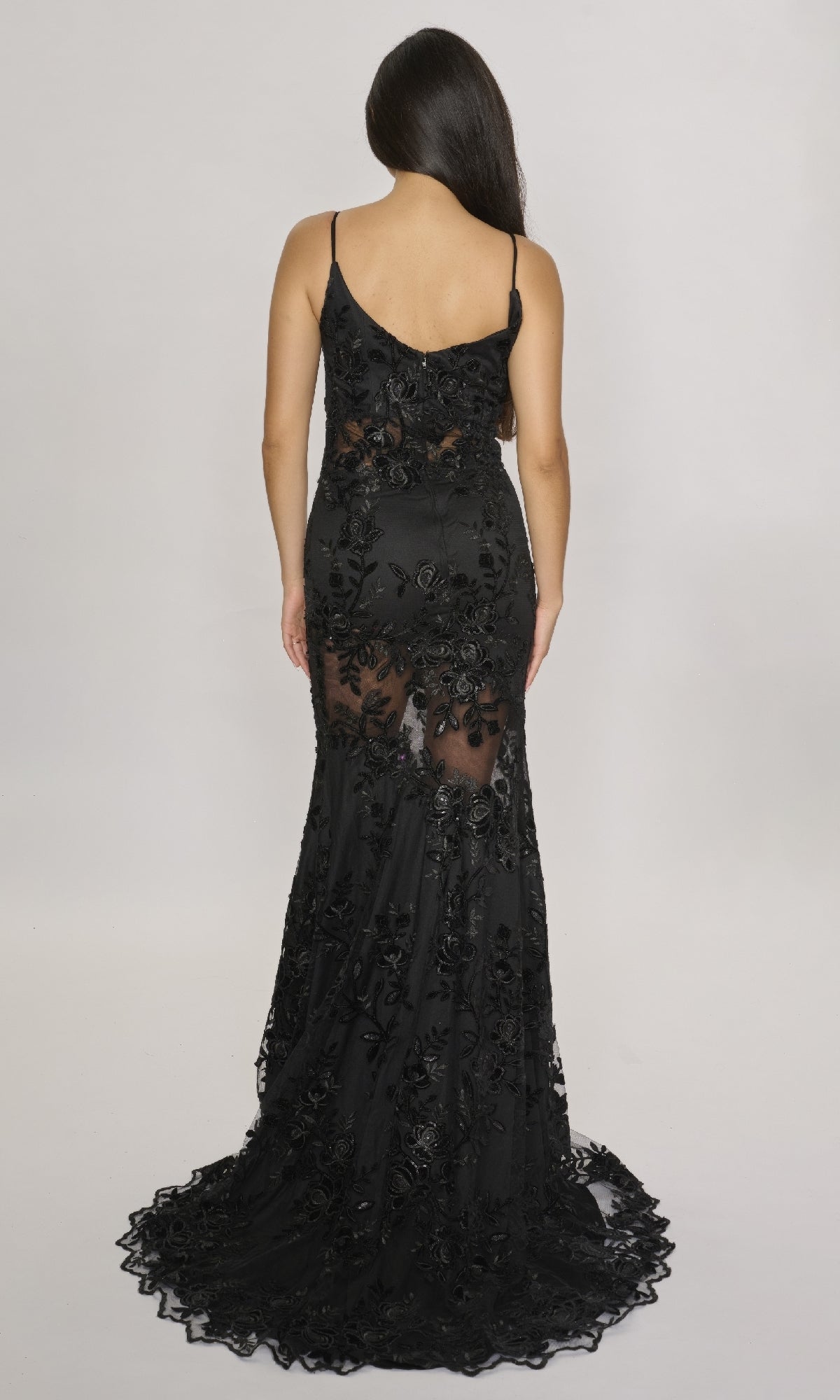 A woman stands facing away, wearing a sleeveless fitted lace-embroidered long dress with sheer floral details, thin straps, and a slight train—the Long Formal Dress: Nina Canacci 2429 in black.