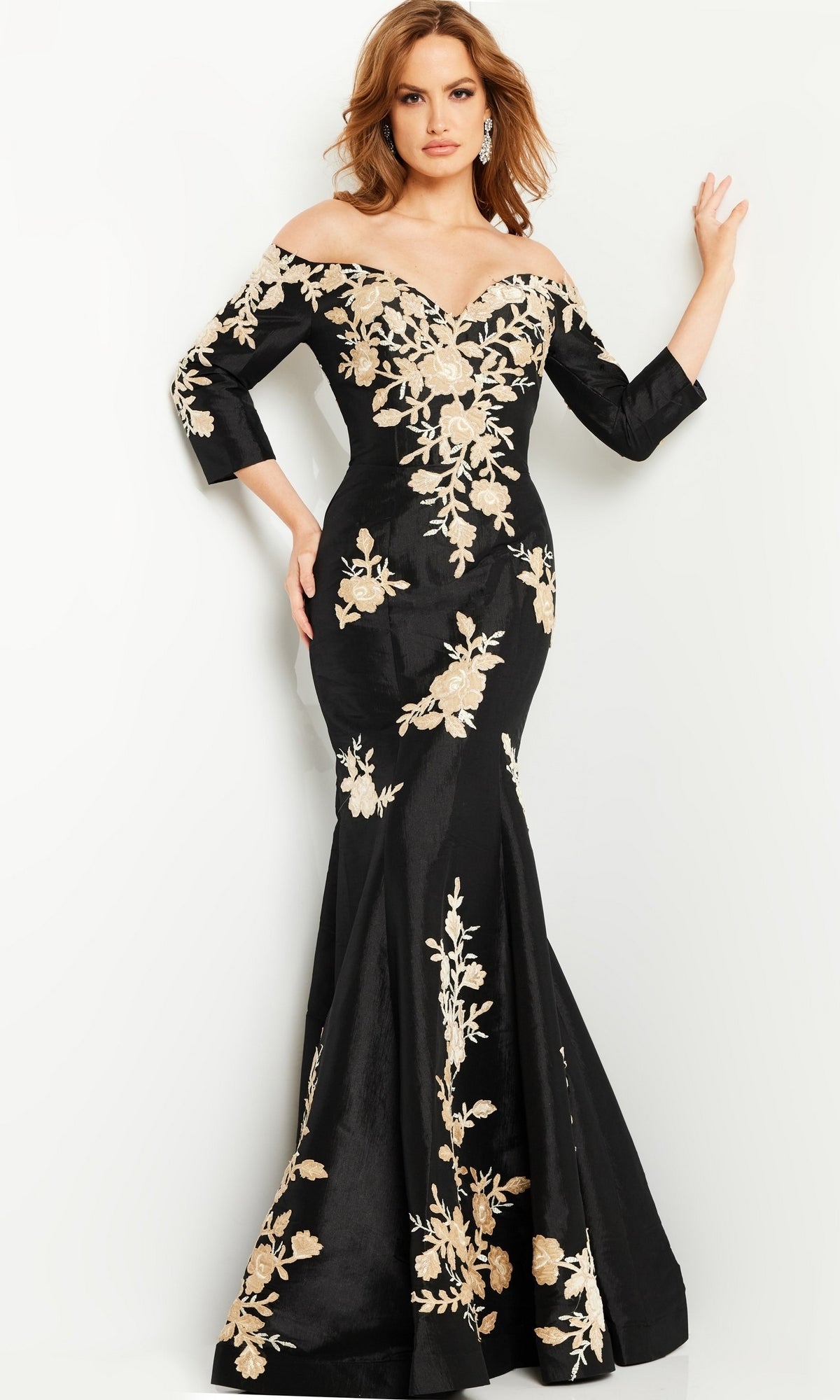 A woman poses against a white background wearing the Black Mermaid Formal Gown 24327, an off-shoulder long black dress with gold floral embroidery.