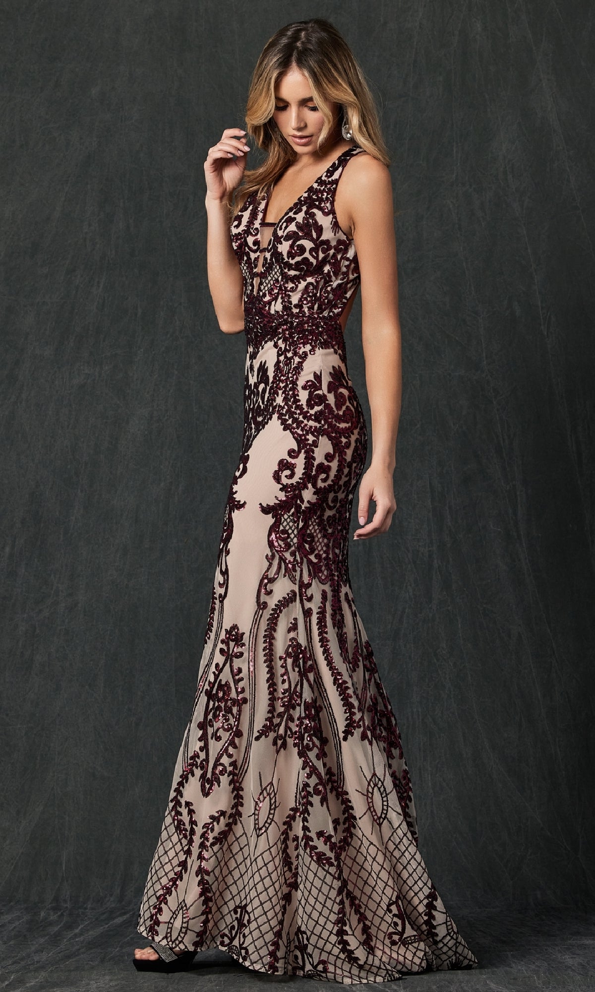 A woman wears the Juliet 243 Long Prom Dress, a form-fitting nude gown with a plunging neckline and detailed burgundy lace, standing against a dark, textured background.