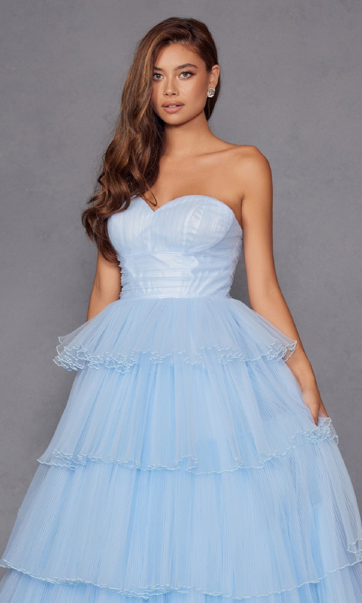 A woman stands against a plain gray background wearing the Strapless Blue Prom Ball Gown: Juliet JT2452K, a long ruffled dress with tiered tulle layers.