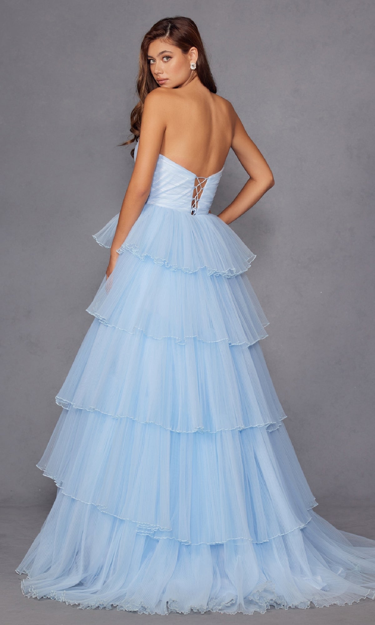 A woman models the Strapless Blue Prom Ball Gown: Juliet JT2452K, featuring ruffles and a lace-up back, against a gray backdrop—an elegant pick for any formal event.