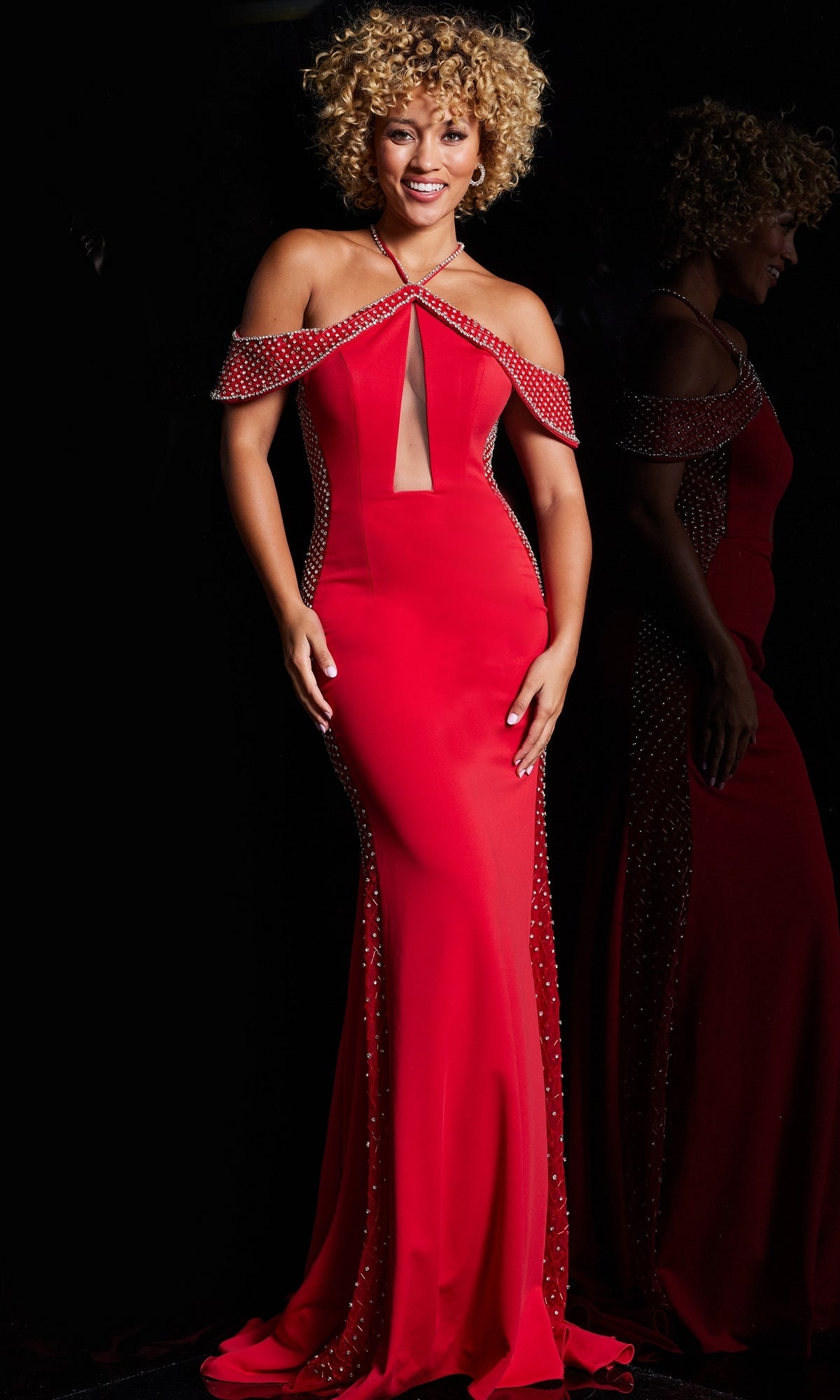 A woman smiles against a dark background wearing the Jovani Sheer-Illusion-Sides Long Prom Dress 24611, a fitted red off-the-shoulder gown with rhinestone embellishments.