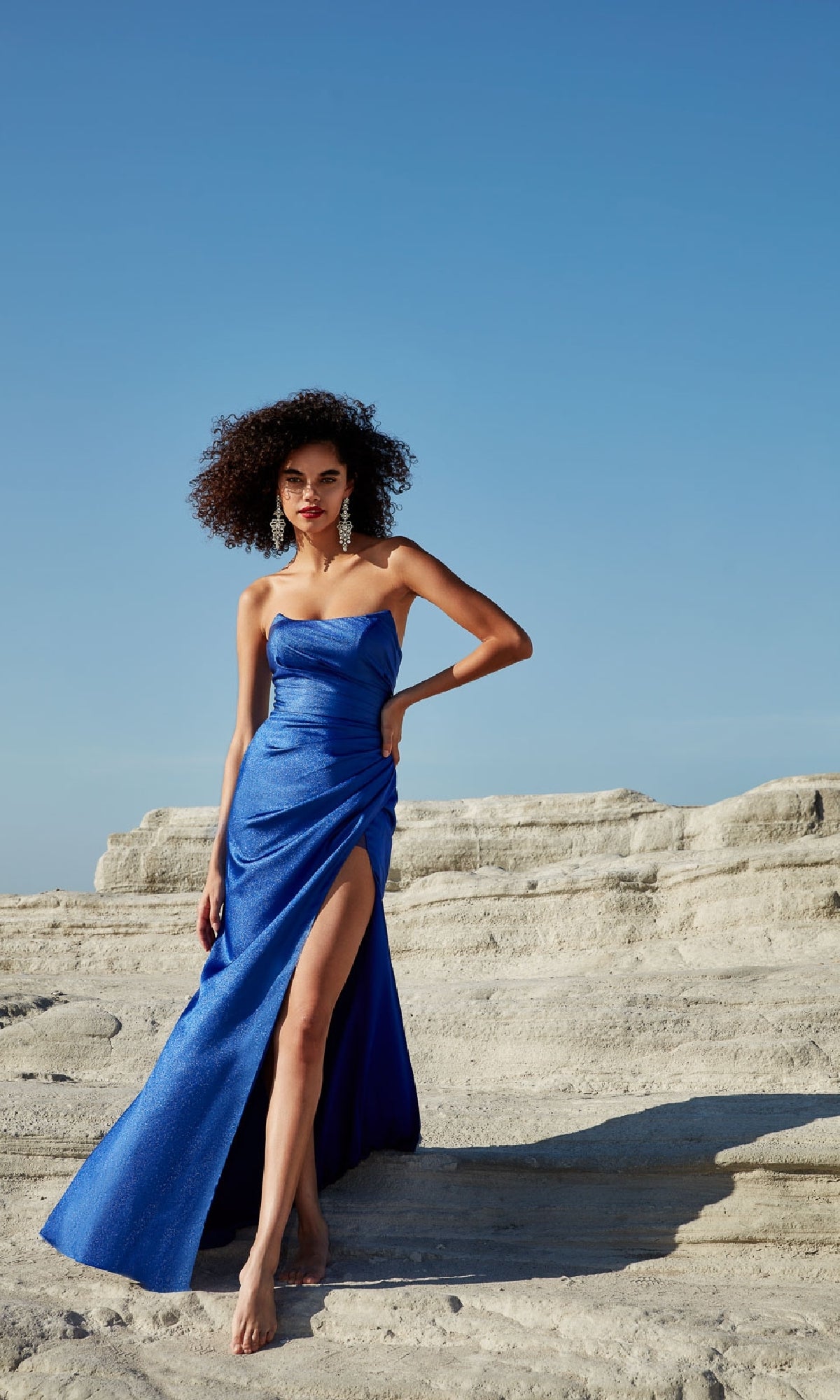 A woman in the Laced Strapless Long Prom Dress: Aerie Couture 247, featuring a thigh-high slit, stands barefoot on rocky terrain beneath a clear blue sky.