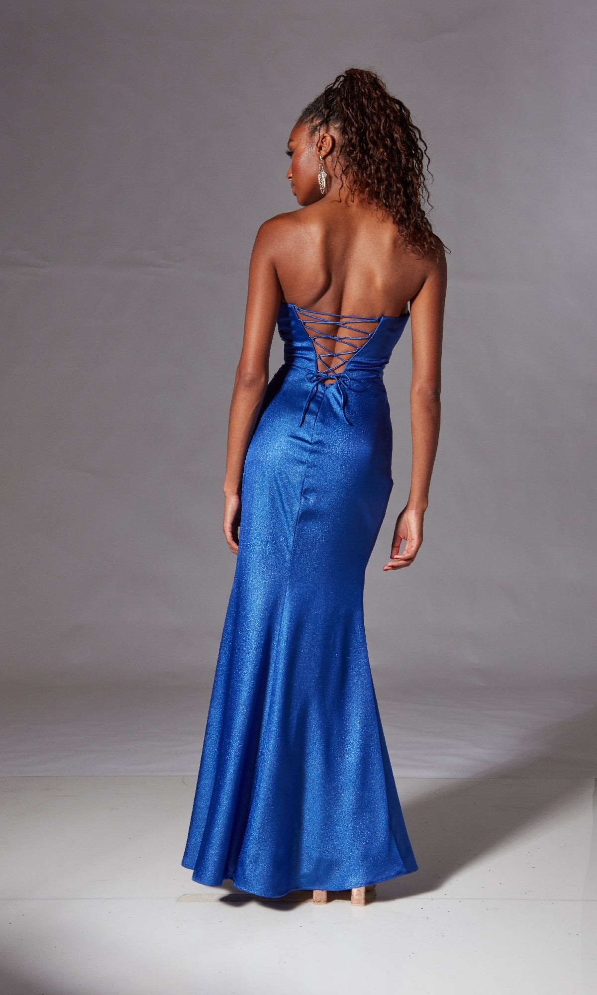 A woman stands with her back to the camera against a plain backdrop, wearing the Laced Strapless Long Prom Dress: Aerie Couture 247 in metallic blue, featuring a floor-length design and lace-up back.