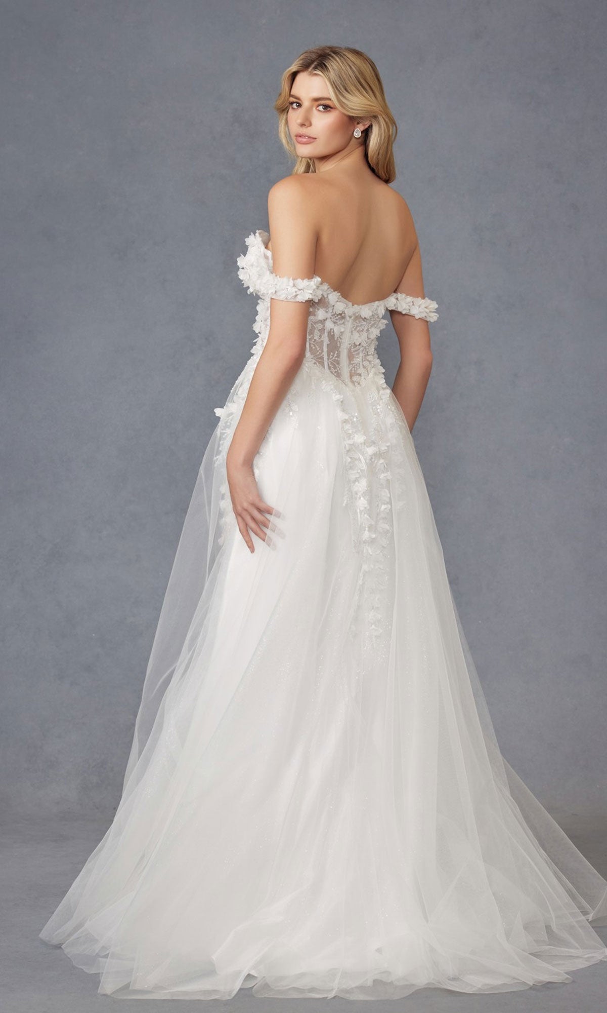 A woman in the Off-White Bridal Ball Gown: Juliet JT2483KW, featuring an off-the-shoulder design, sheer back, floral details, lace accents, and an embroidered corset bodice, stands against a plain gray background looking over her shoulder.