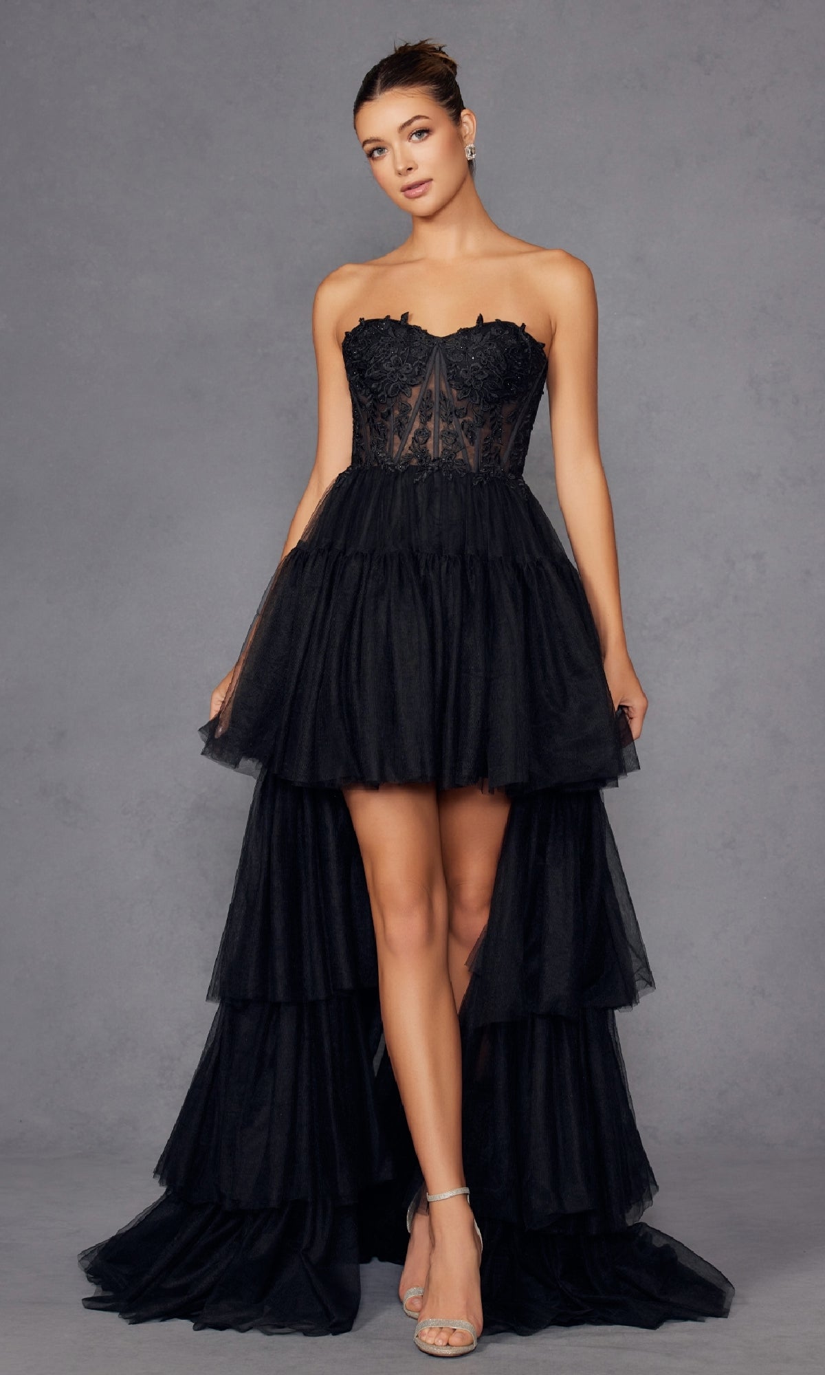 A woman in the Juliet JT2488S High-Low Strapless Prom Dress, featuring a lace corset bodice and layered ruffled skirt, pairs it with silver heels while standing against a plain gray background.