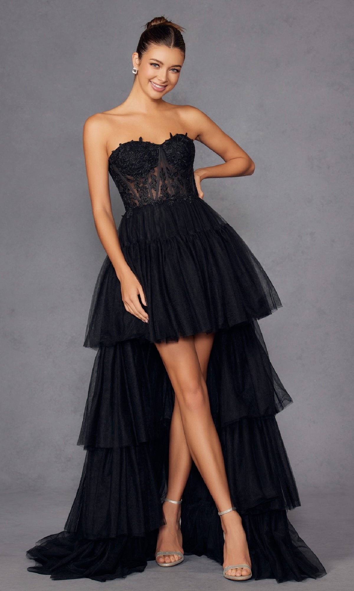 A woman models the High-Low Strapless Prom Dress: Juliet JT2488S, showcasing its lace corset bodice and tiered tulle skirt against a plain gray background.
