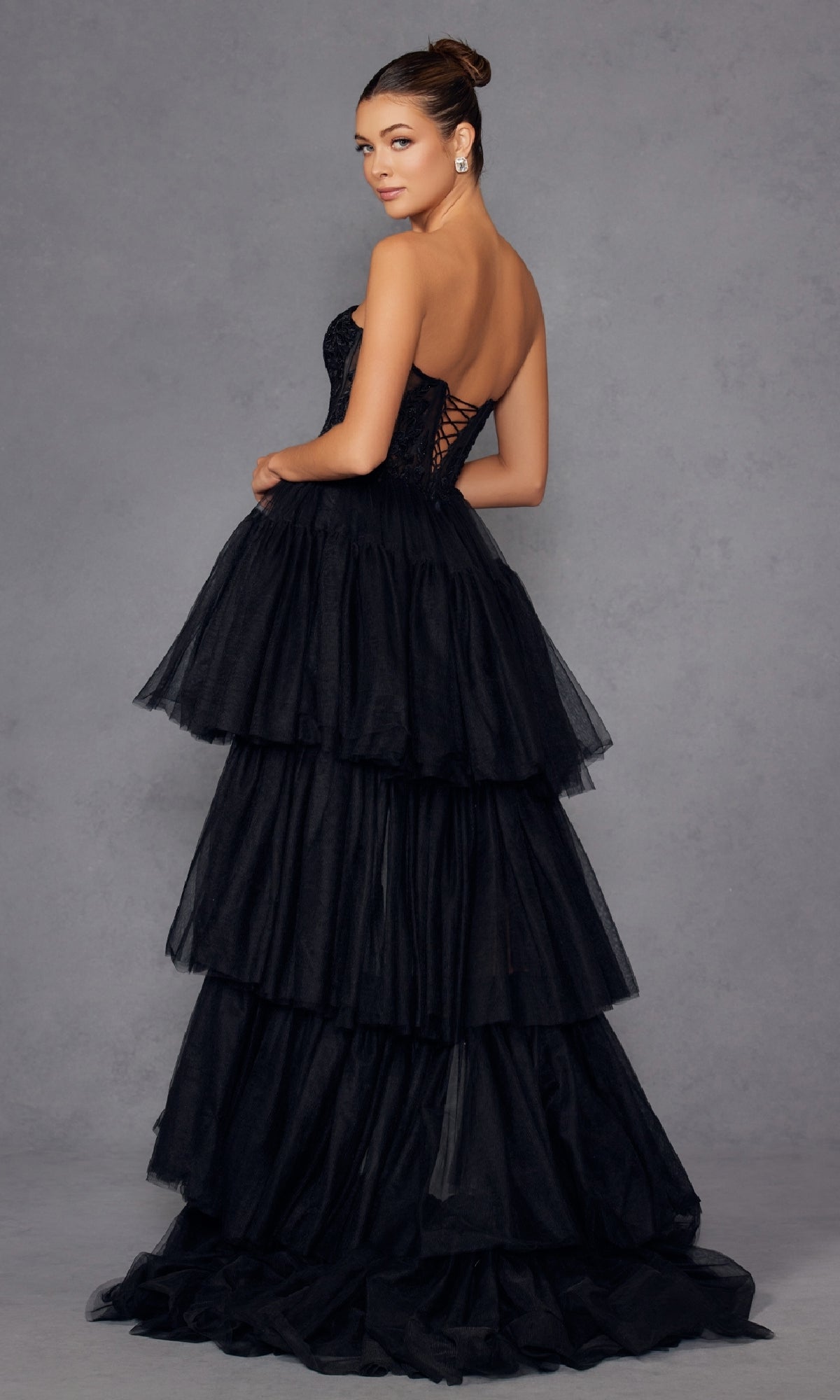 A woman models the Juliet JT2488S High-Low Strapless Prom Dress with a lace corset bodice and removable floor-length skirt, standing against a plain gray background and looking over her shoulder.
