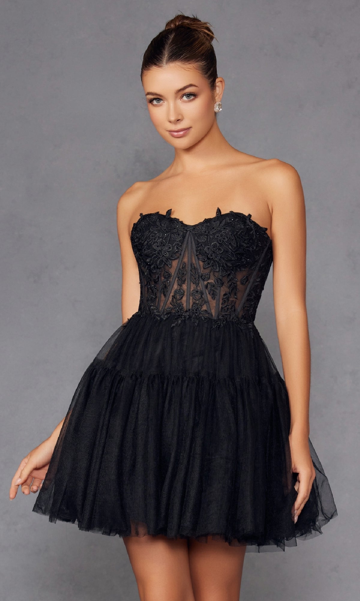 A woman stands against a plain gray background wearing the High-Low Strapless Prom Dress: Juliet JT2488S, featuring a lace corset bodice and flared tulle skirt.
