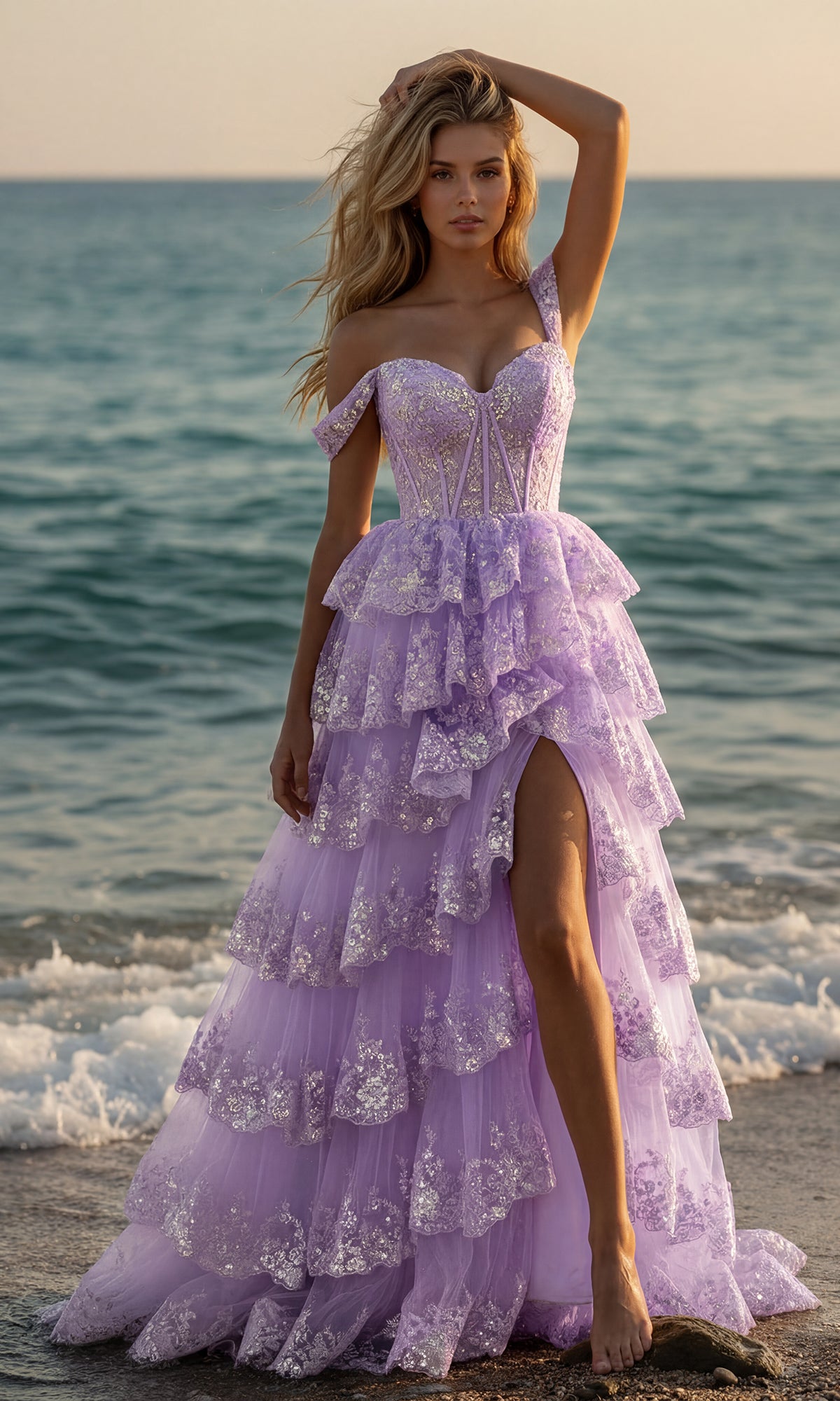 A woman stands on the beach in the PromGirl 250005 A-Line Corset Lace Prom Dress, a lilac gown featuring ruffled tiers, sequin details, an off-the-shoulder design, and a thigh-high slit with the ocean as a stunning backdrop.