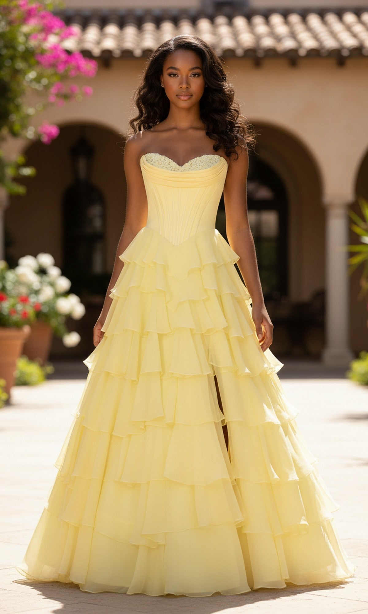 A woman stands outdoors in front of a building, wearing the PromGirl 250057—a strapless yellow ruffled prom ball gown with elegant layered tiers.