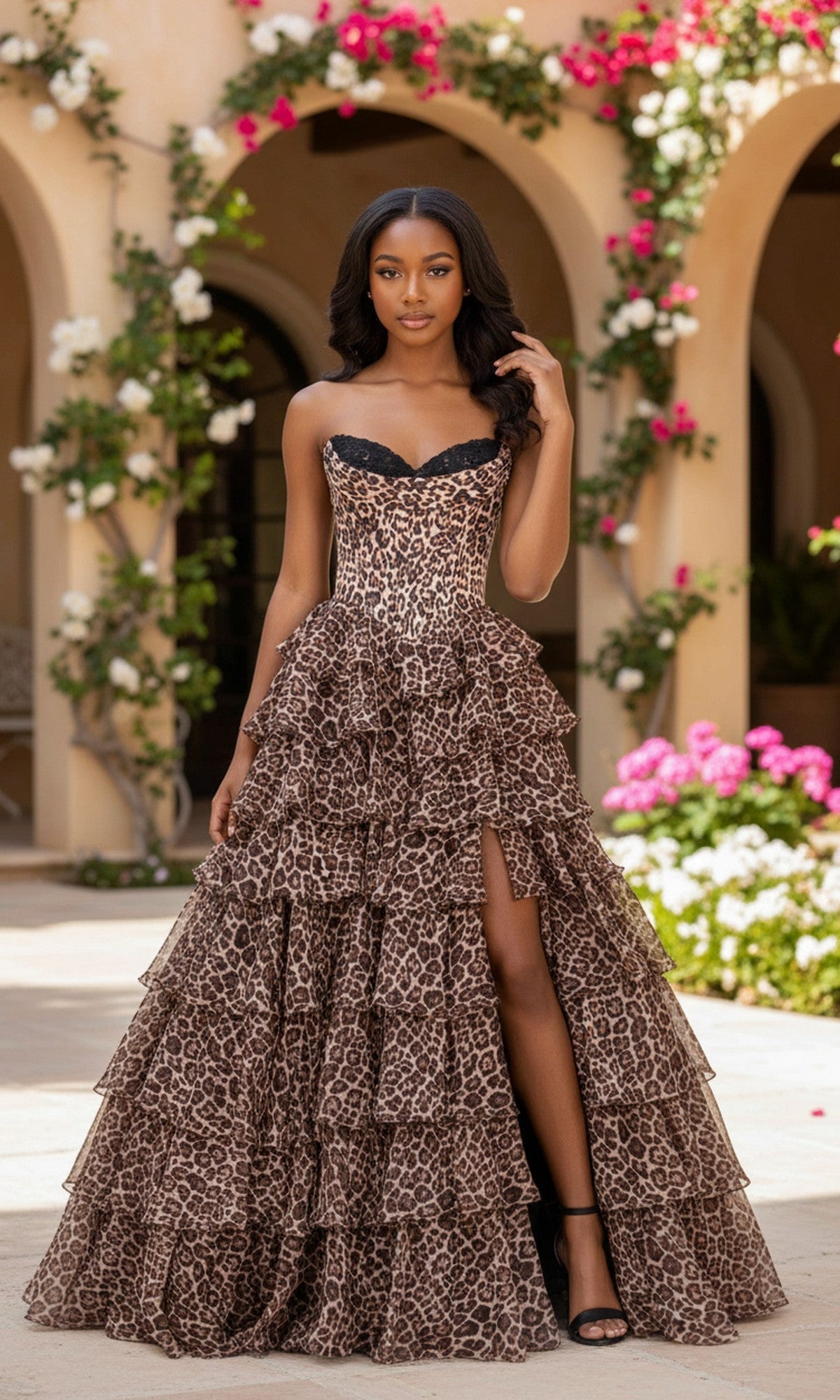 A woman stands outdoors in front of a vine-covered building, wearing the Leopard Long Prom Gown: PromGirl Exclusive 250123A with a strapless neckline, chiffon a-line skirt, tiered layers, thigh-high slit, and black heels.
