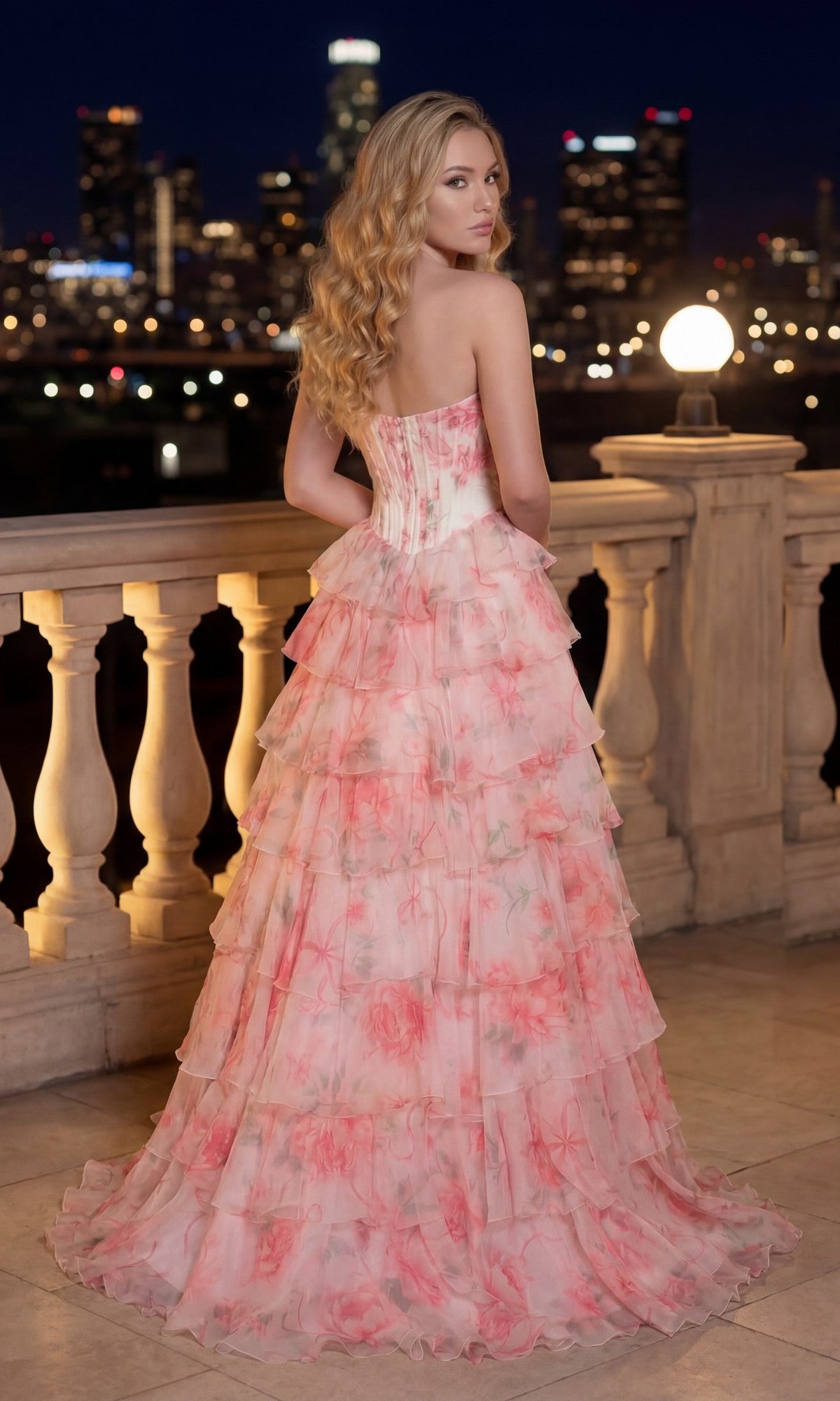 A woman with wavy blonde hair wears the Floral Prom Ball Gown: PromGirl Exclusive 250123D, a strapless pink floral-print dress, while standing on a balcony at night with city lights twinkling behind her.