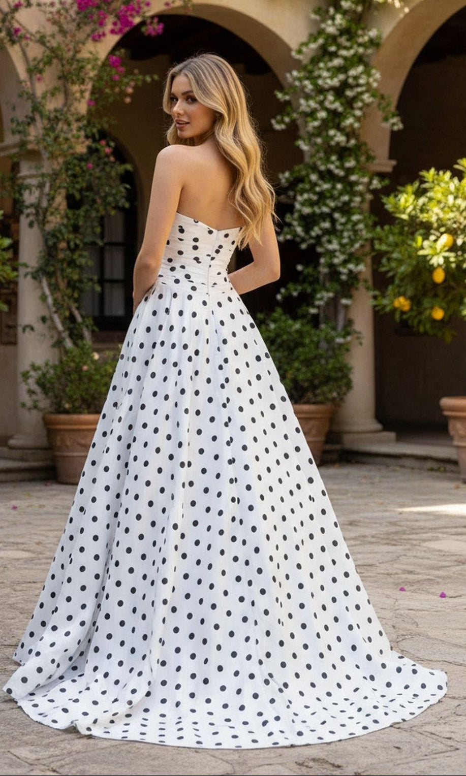 A woman stands outdoors in the PromGirl Exclusive 250209 long prom dress, its strapless, floor-length A-line silhouette flowing gracefully as she looks over her shoulder amid potted plants and arches.