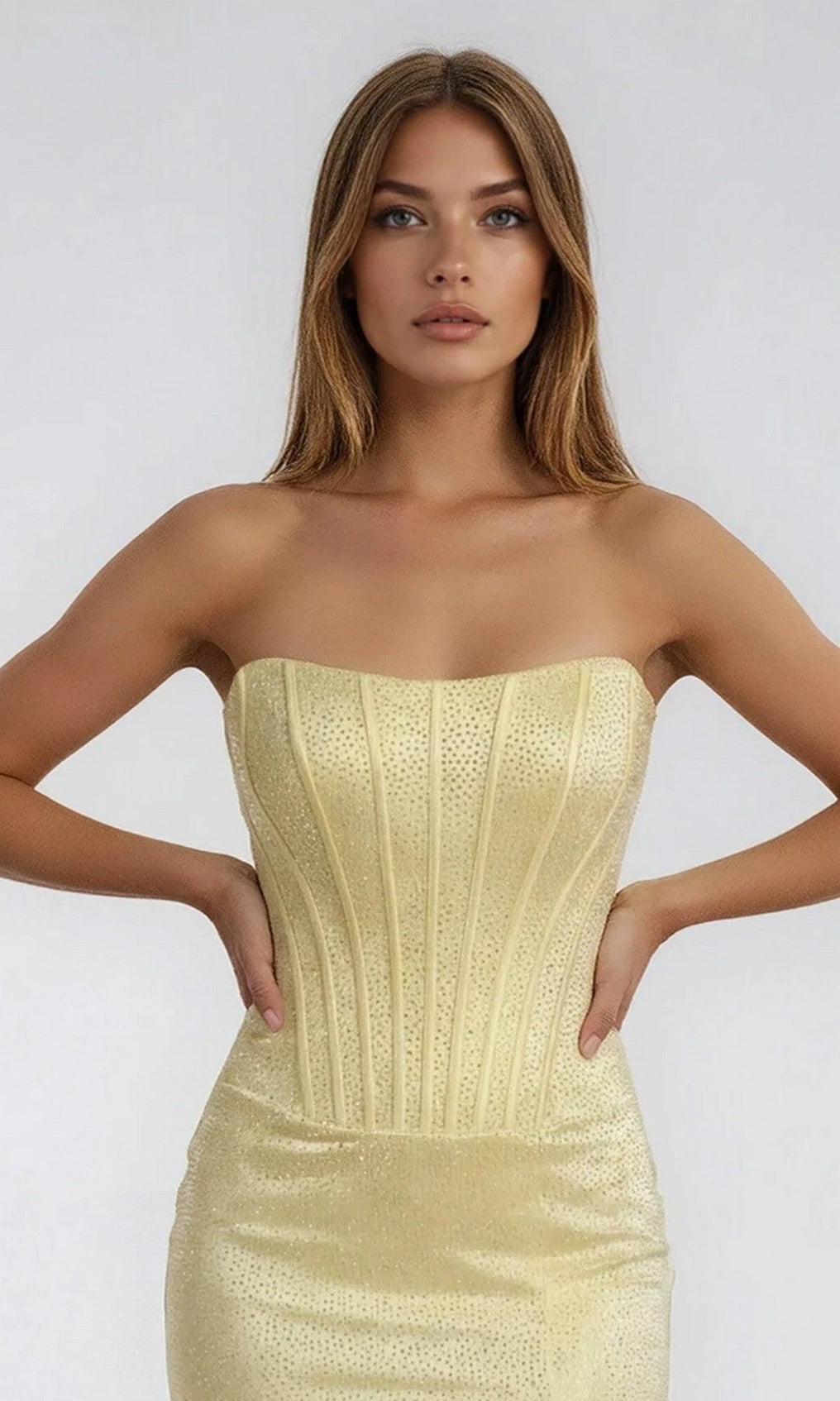 A woman with straight light brown hair models the PromGirl Exclusive 250230, a strapless, fitted pale gold long stretch satin prom dress featuring a textured pattern and subtle leg slit, standing against a plain light background.