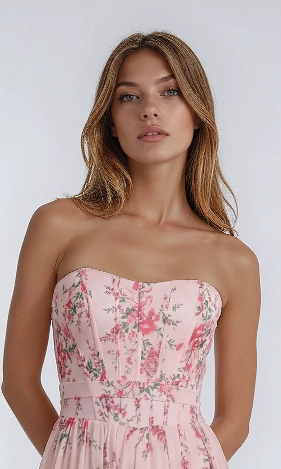 A woman poses against a plain background wearing the PromGirl Exclusive 250236, a long strapless prom dress in a light pink hue with an elegant floral pattern.