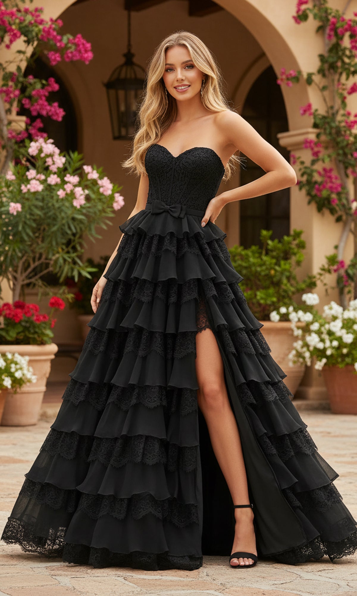 A woman stands outdoors on a stone patio with potted plants, wearing the PromGirl Exclusive 250238—an elegant long black strapless prom dress featuring tiered layers, a lace corset bodice, and a front slit.