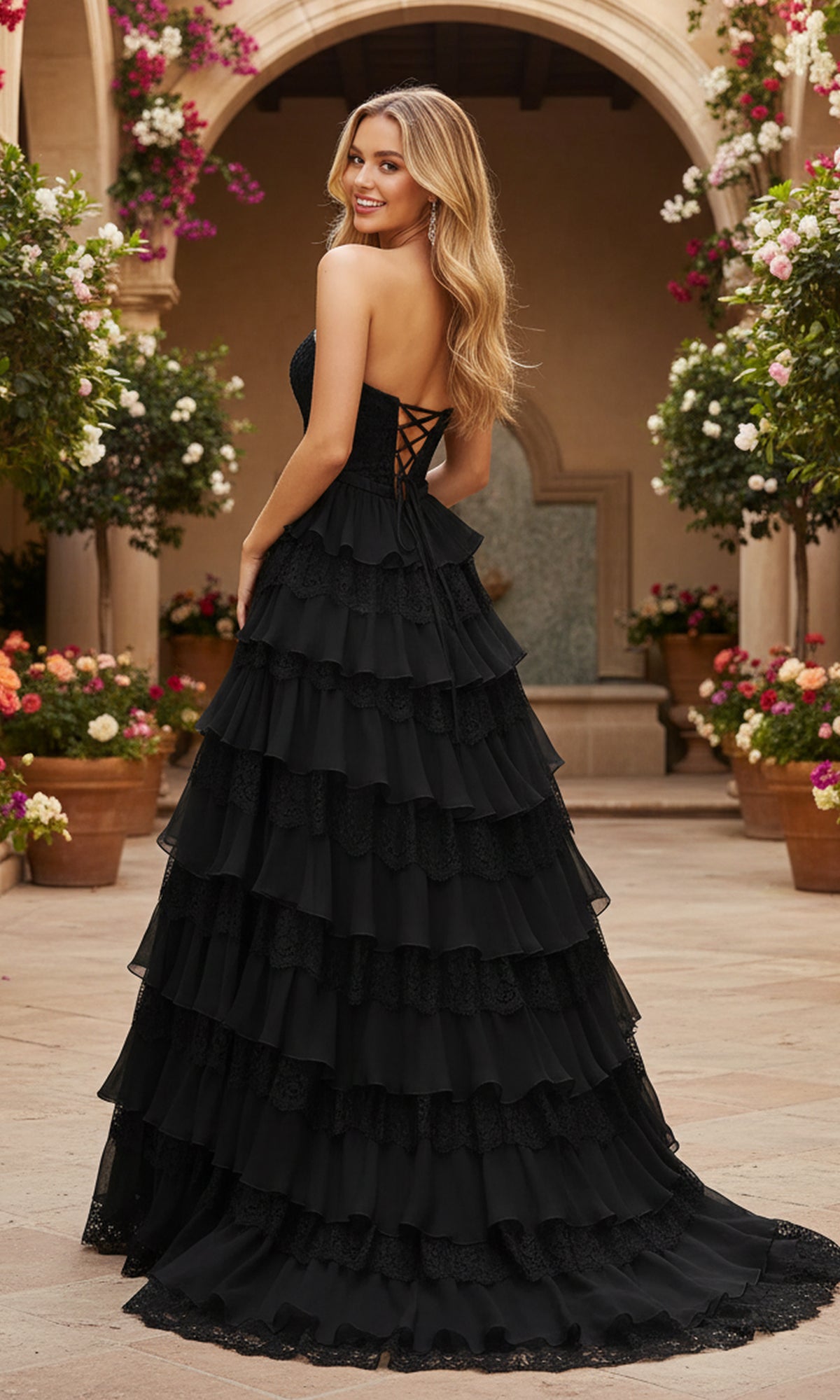 A woman wearing the PromGirl Exclusive 250238 long black strapless lace prom dress stands outdoors on a stone patio among potted flowers, glancing back over her shoulder at the camera.