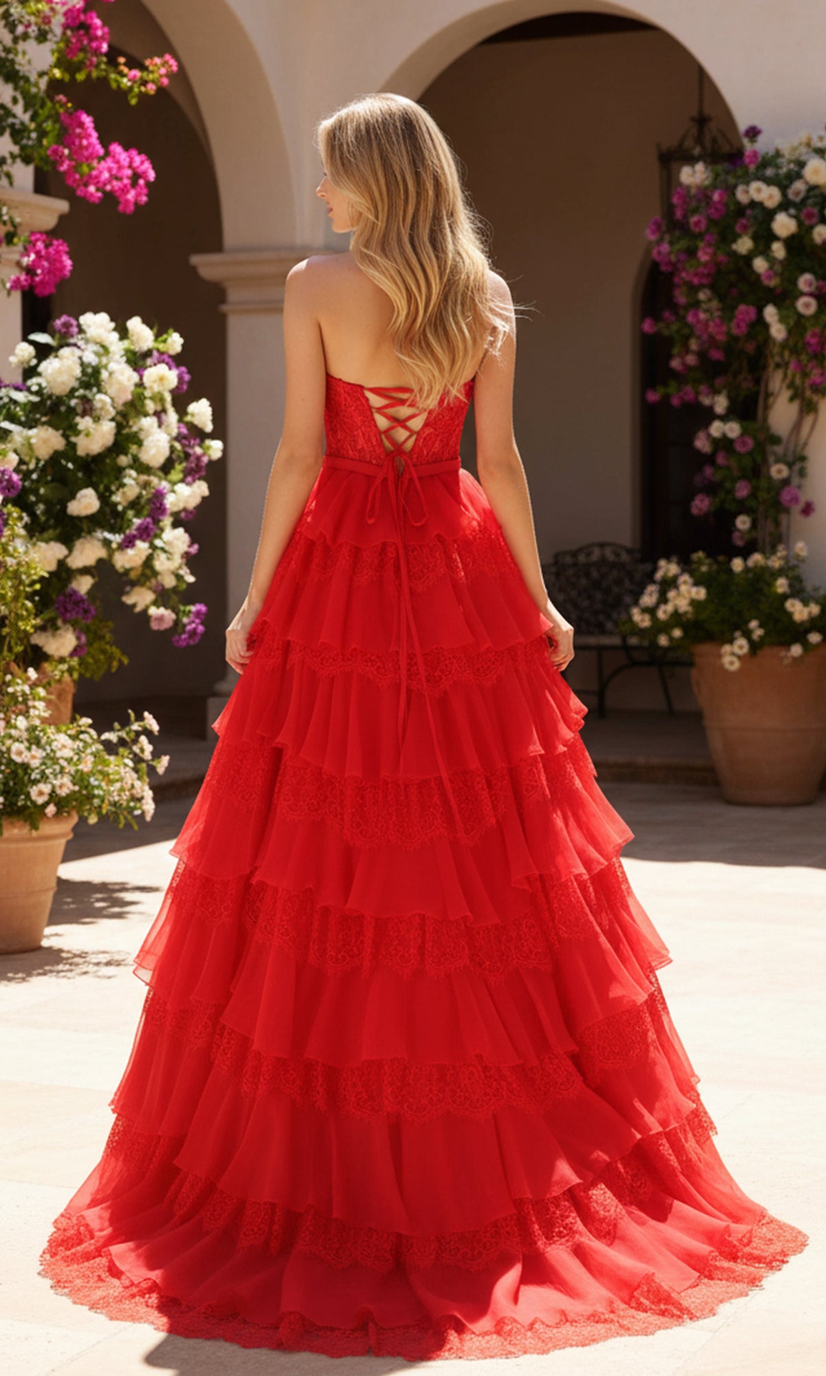 A woman with long blonde hair wears the Long Prom Dress: PromGirl Exclusive 250238, a strapless tiered red gown with a lace-up back, standing outdoors among potted flowers and arched architecture.