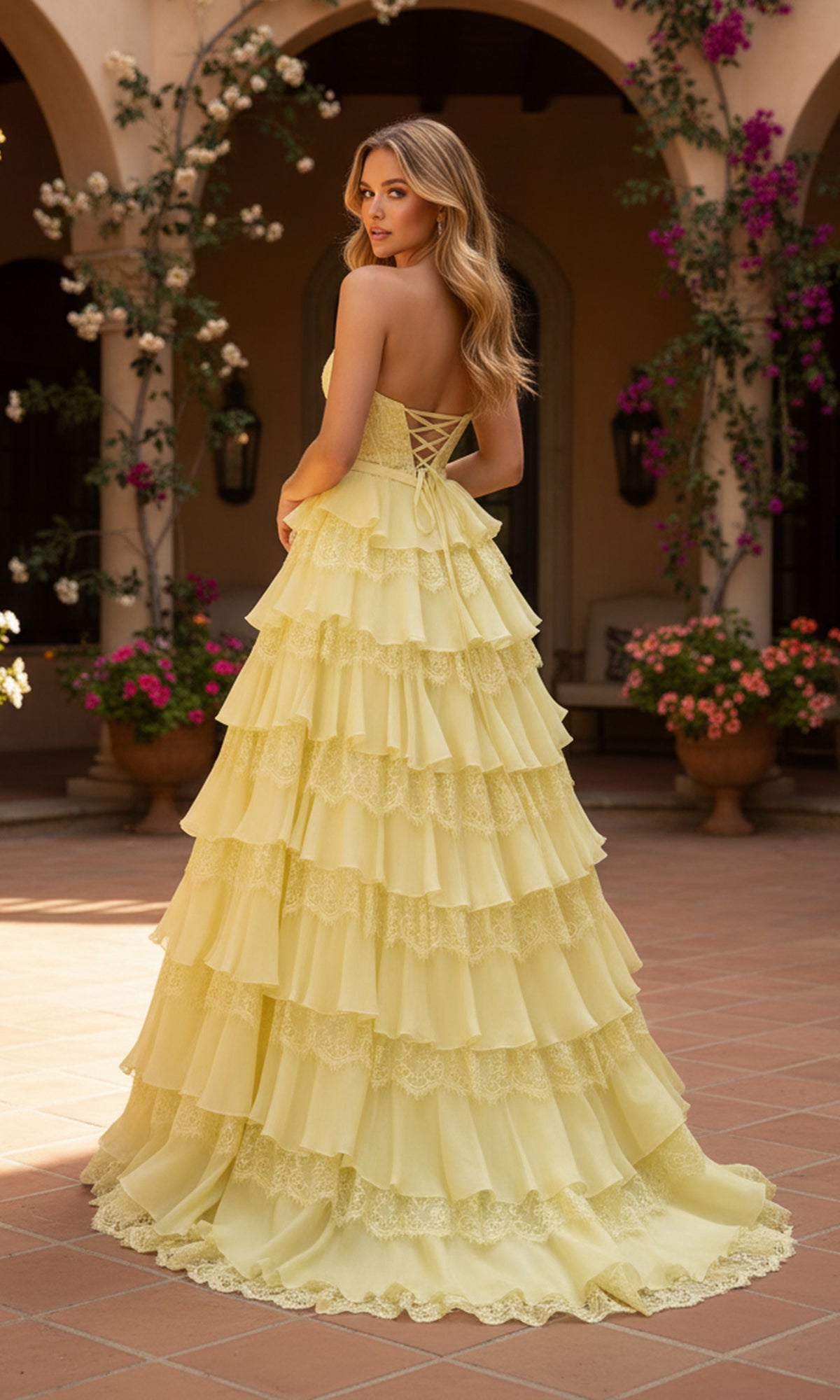 A woman in the PromGirl Exclusive 250238, a strapless yellow tiered ruffle ball gown, stands outside on a tiled patio with flowering plants and arched doorways.