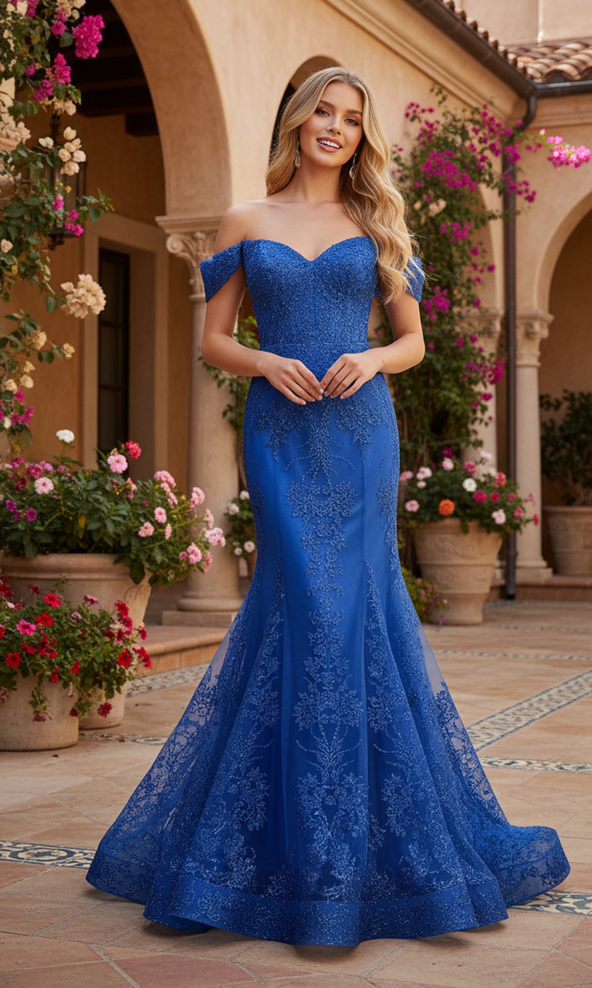 A woman wearing the PromGirl Exclusive 250250, a long off-the-shoulder blue lace prom dress, stands in a courtyard with potted flowers and arched architecture in the background.