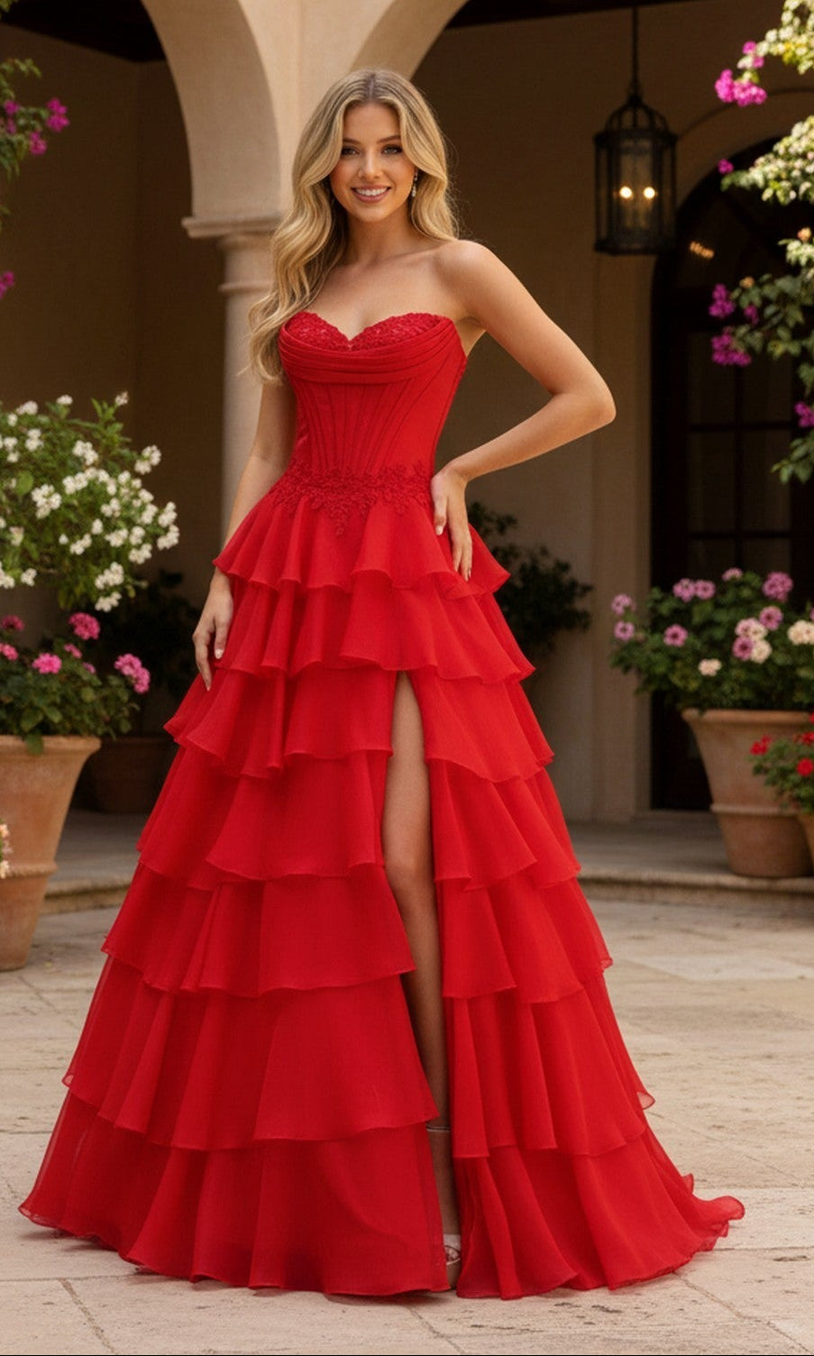 Outdoors, a woman wears the Long Prom Dress: PromGirl Exclusive 250267, which features a strapless corset bodice and a tiered ruffled skirt with a front slit. She stands among potted flowers with an arched building in the background.