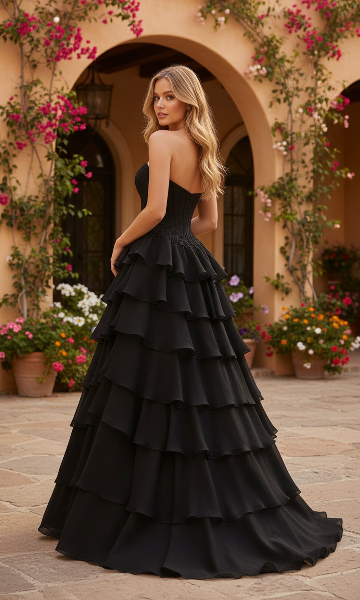 A woman in the PromGirl Exclusive 250267 long prom dress with a strapless sweetheart neckline and corset bodice stands in a courtyard filled with flowers, framed by an arched building.