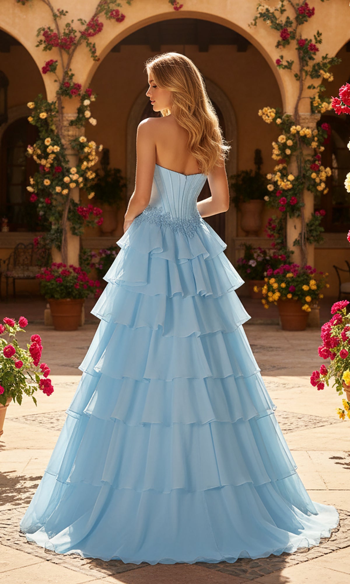 A woman with long blonde hair wears the PromGirl Exclusive 250267 long prom dress in light blue, standing in a sunny courtyard filled with potted flowers and surrounded by arched walls.