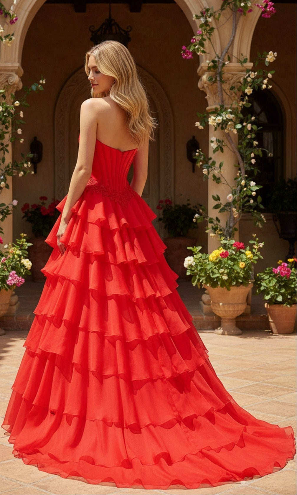A blonde woman stands outdoors among potted plants and flowers wearing the PromGirl Exclusive 250267, a long strapless prom gown with a corset bodice and tiered ruffled skirt.