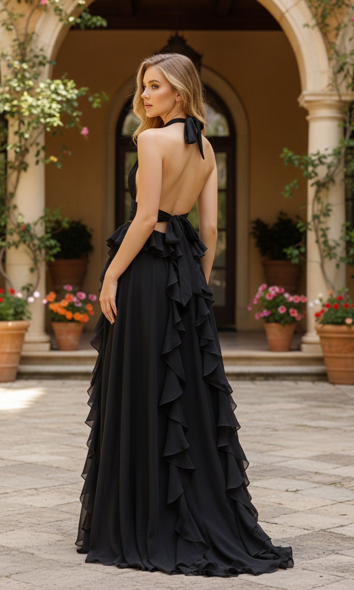 A woman wearing the PromGirl Exclusive 250269—a floor-length black chiffon prom dress with a halter neckline and open back—stands outdoors in front of an arched doorway and potted plants.
