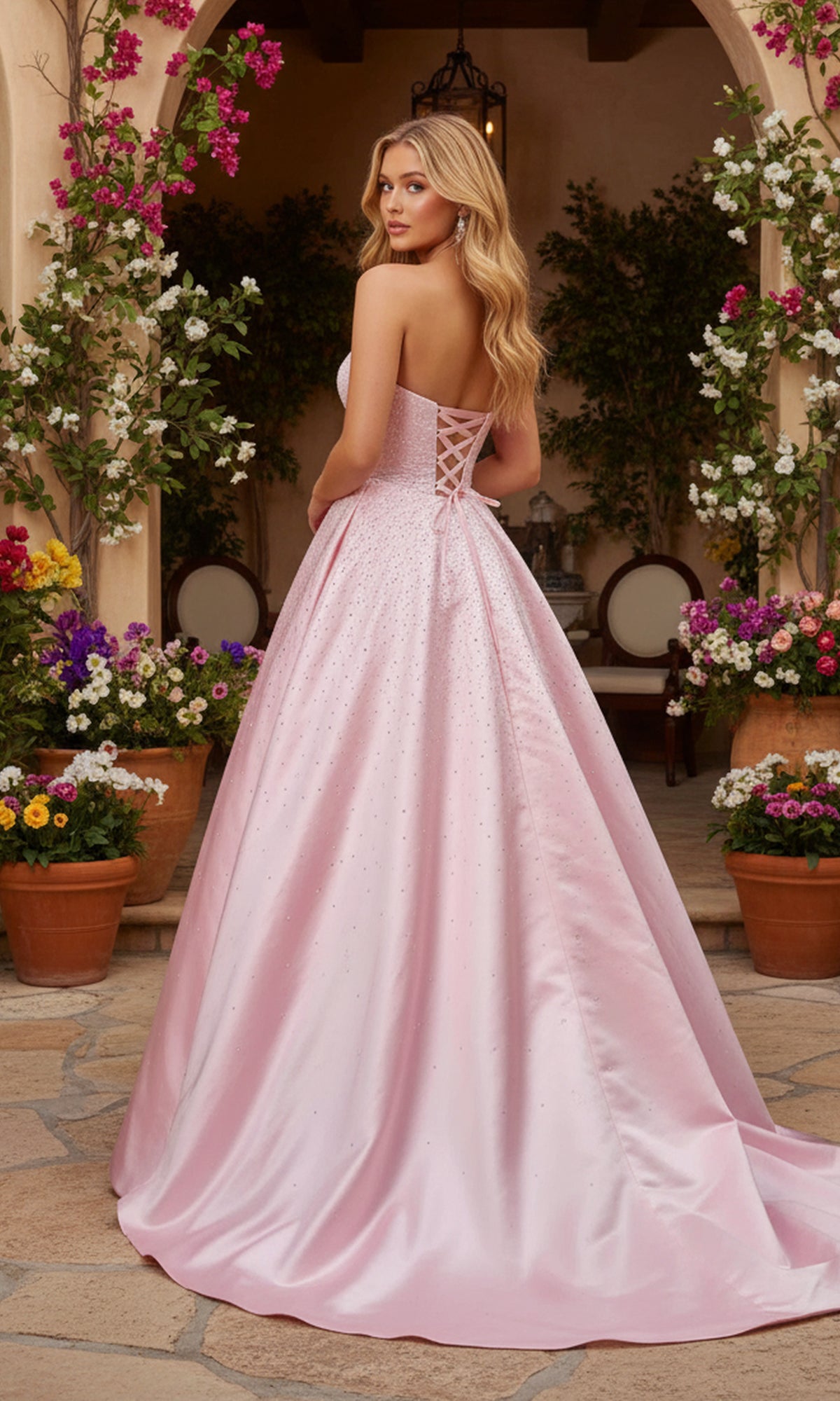 Wearing the PromGirl Exclusive 250297 long prom dress, a woman stands outdoors among potted flowers, her strapless satin gown with a lace-up back shimmering with rhinestone pearl embellishments.