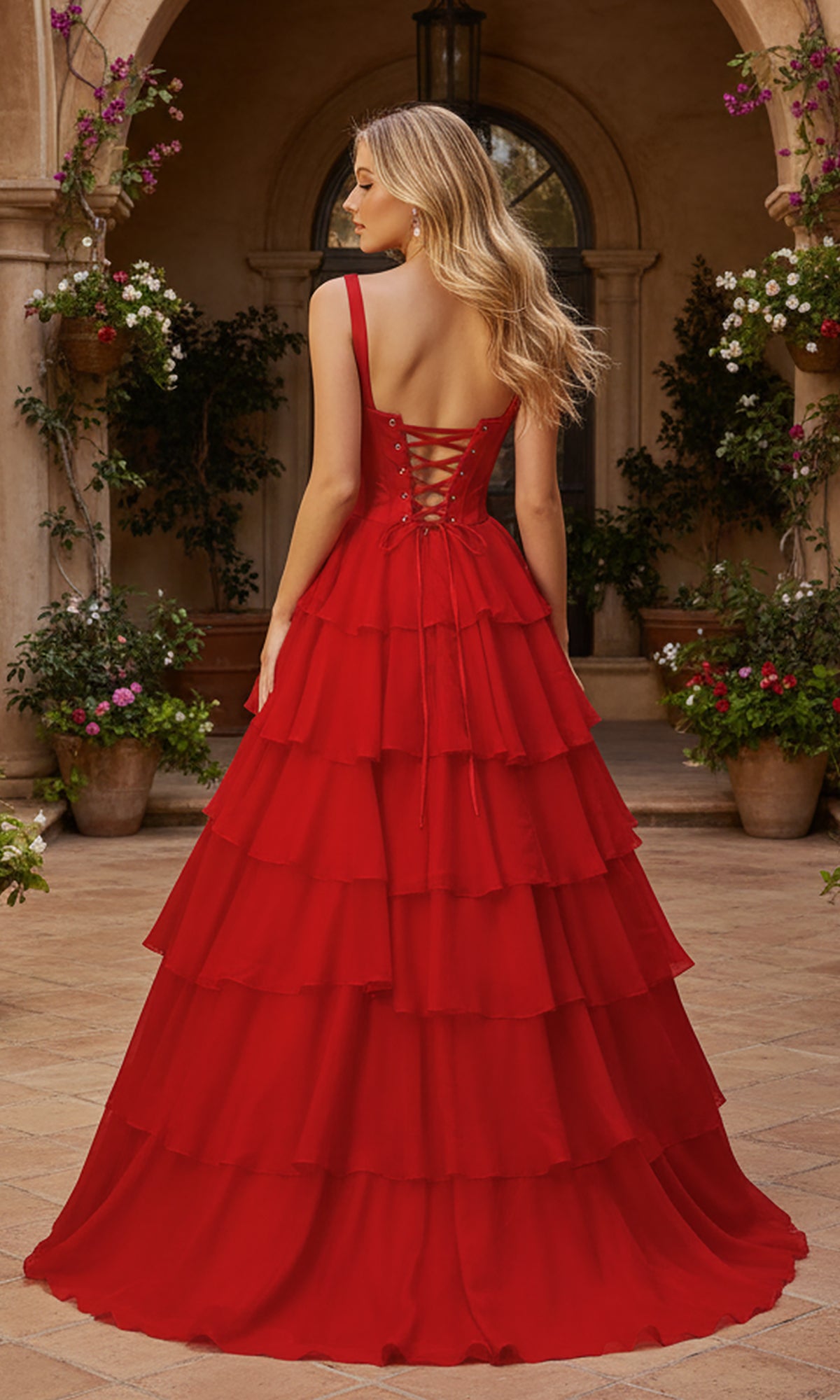 Wearing the PromGirl Exclusive 250300, a layered floor-length red prom ball gown with a lace-up back, a woman stands outdoors among potted plants and an arched doorway.