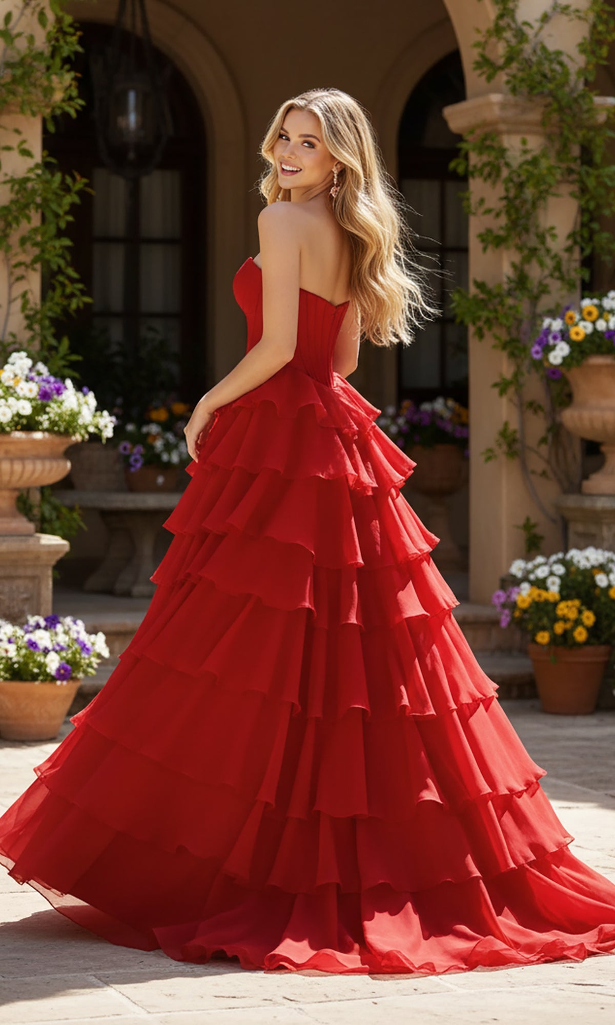 A woman poses outdoors in front of a building, smiling over her shoulder while wearing the PromGirl Exclusive 250302 long prom dress featuring a strapless, ruffled gown with a layered skirt, surrounded by potted flowers.
