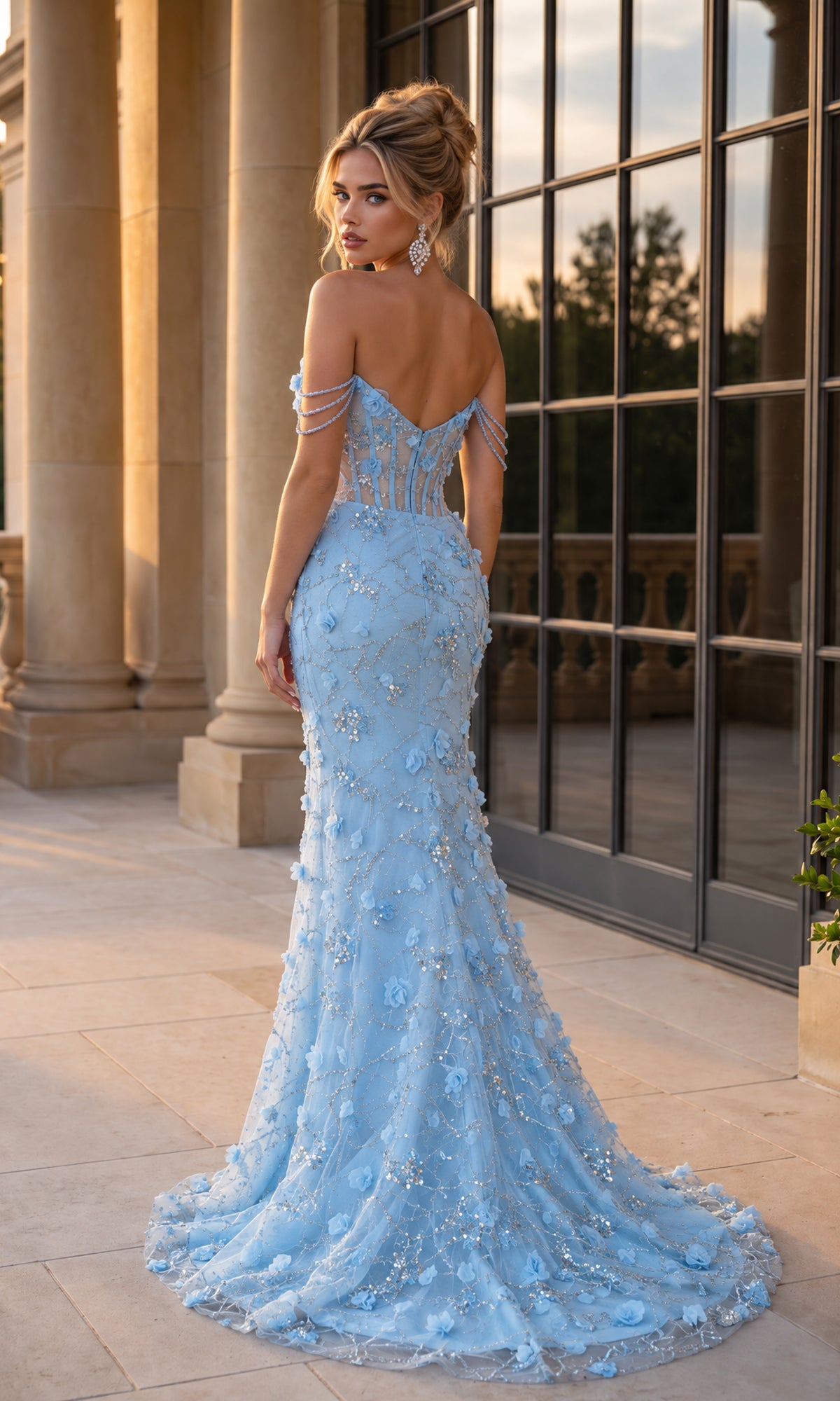 Wearing the PromGirl 250356 Off-Shoulder 3D Floral Prom Dress, a woman stands on a stone terrace before tall windows and columns at sunset. The light blue, floor-length gown features beaded straps, delicate 3D floral appliqué, and a sheer corset bodice that enhances her elegant silhouette with an ethereal touch.