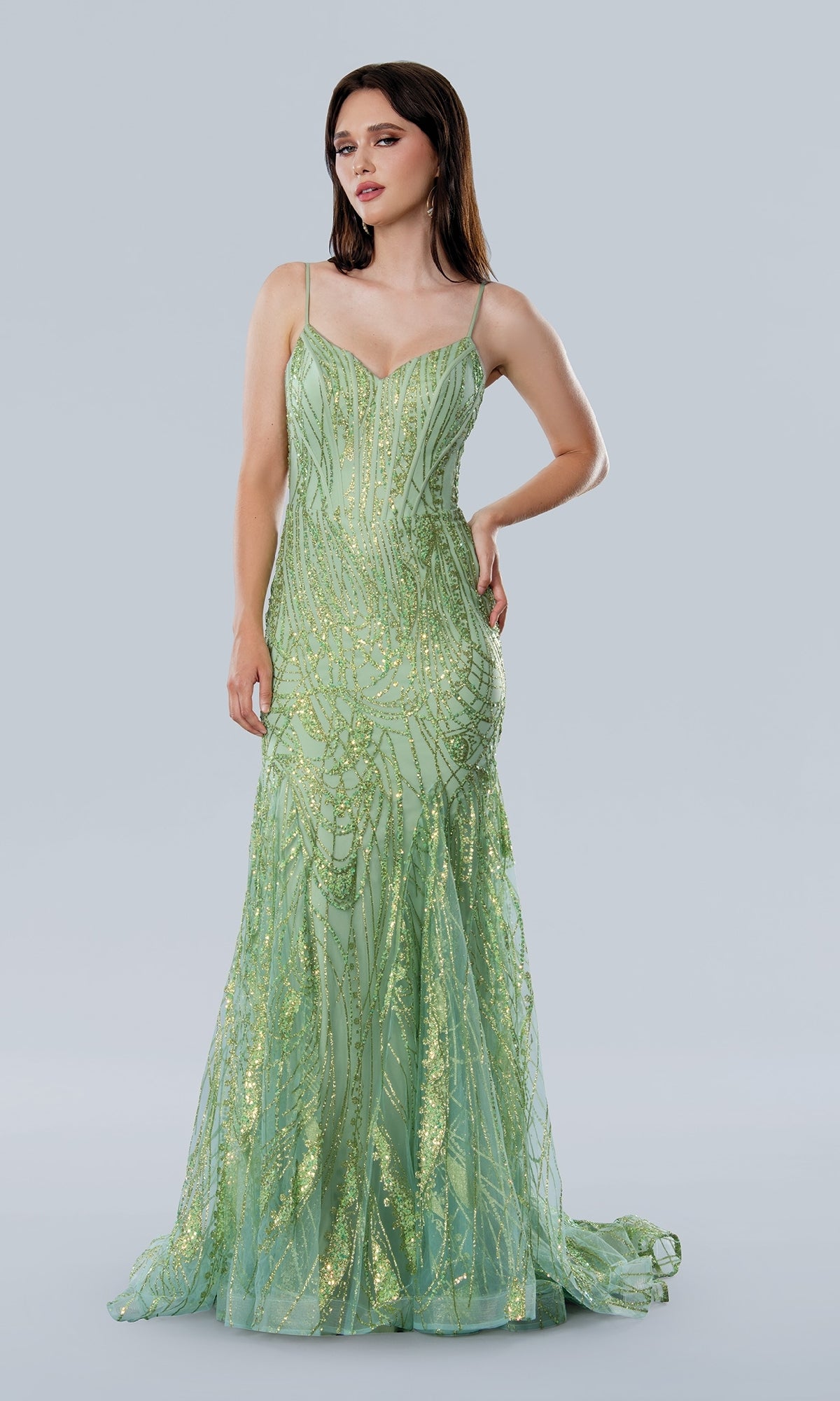A woman wears the Stella Couture 25105, a floor-length light green prom dress with thin straps, a corset bodice, and gold sequin details, standing against a plain gray background.
