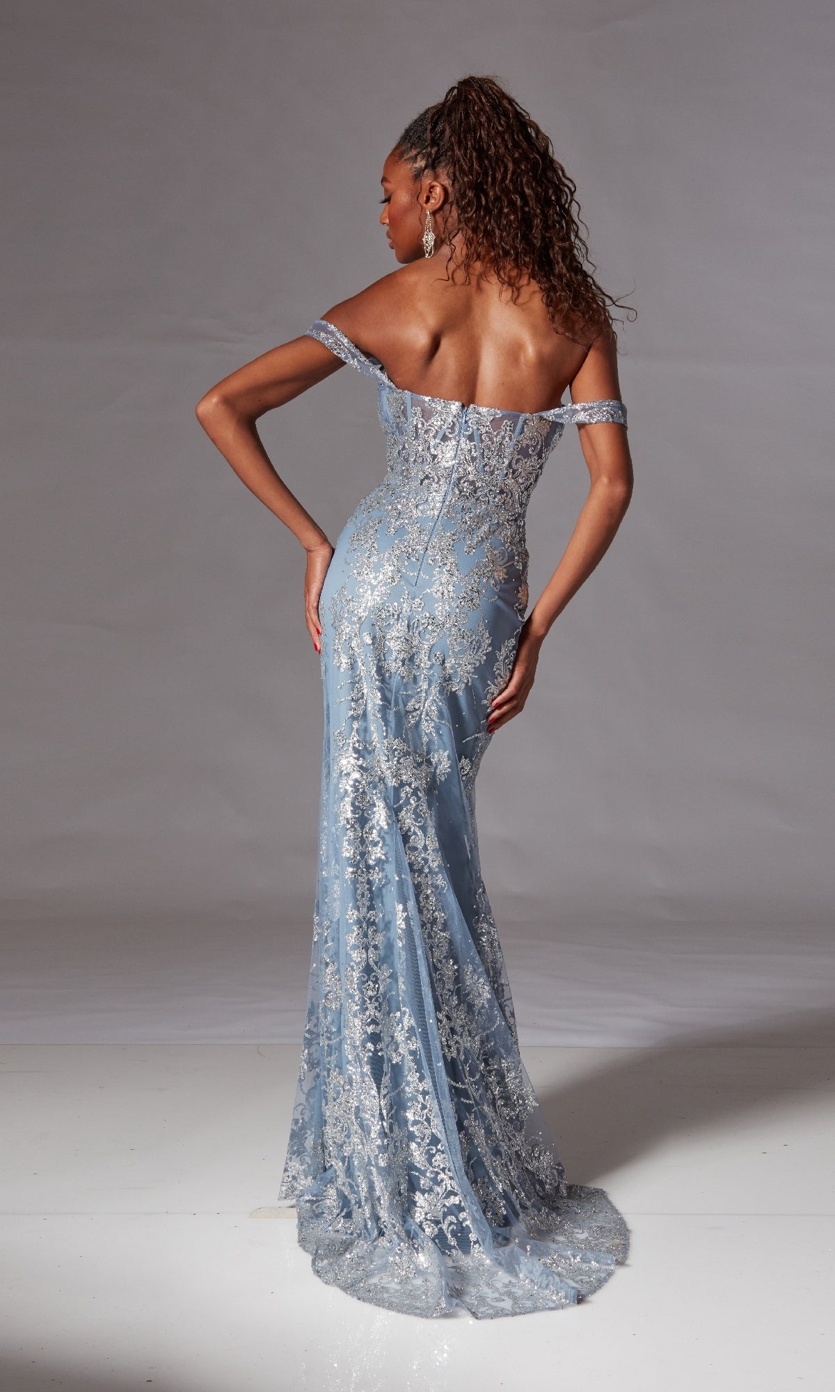 A woman poses facing away from the camera in the Long Blue Glitter Prom Dress: Aerie Couture 252, featuring off-the-shoulder style and silver lace detailing, set against a neutral background.