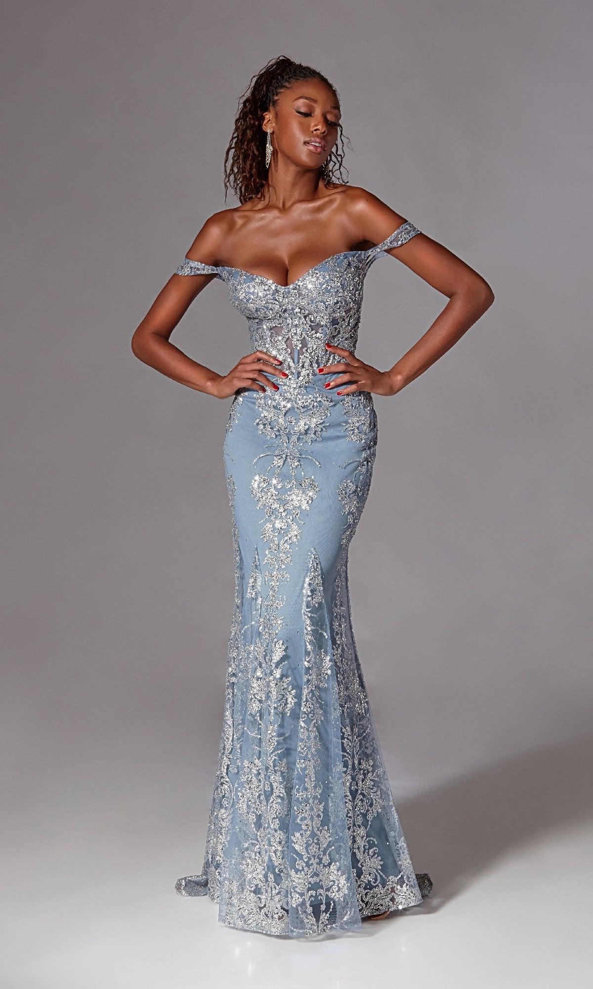 A woman stands confidently with hands on hips, wearing the Aerie Couture 252 Long Blue Glitter Prom Dress—a fitted light blue evening gown with silver lace and beading—against a plain backdrop.