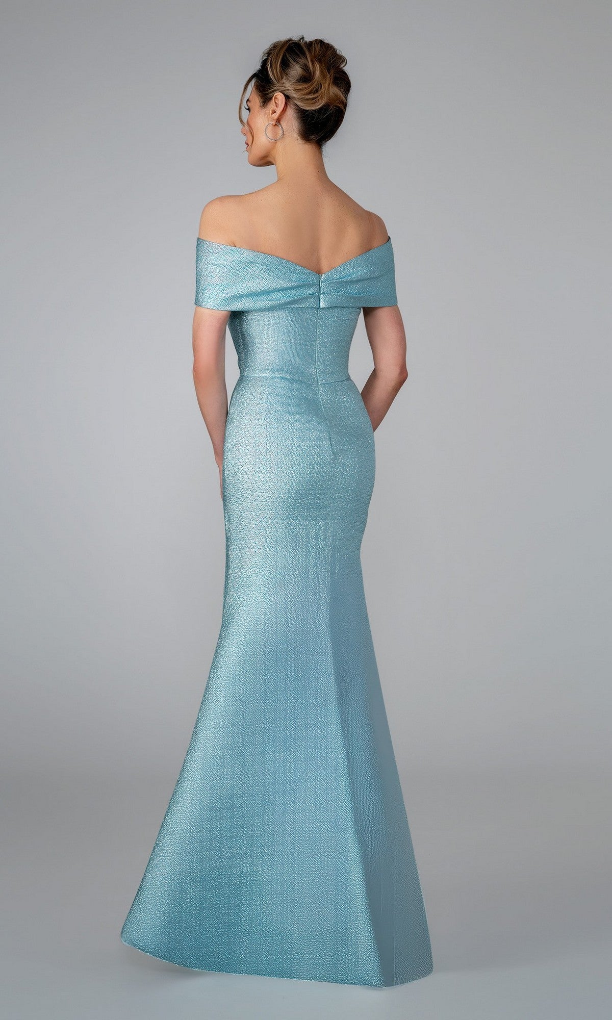A woman in the Stella Couture 25329 long formal dress—an off-the-shoulder, fitted, light blue mermaid gown with a subtle sheen—stands with her back to the camera against a plain grey backdrop.