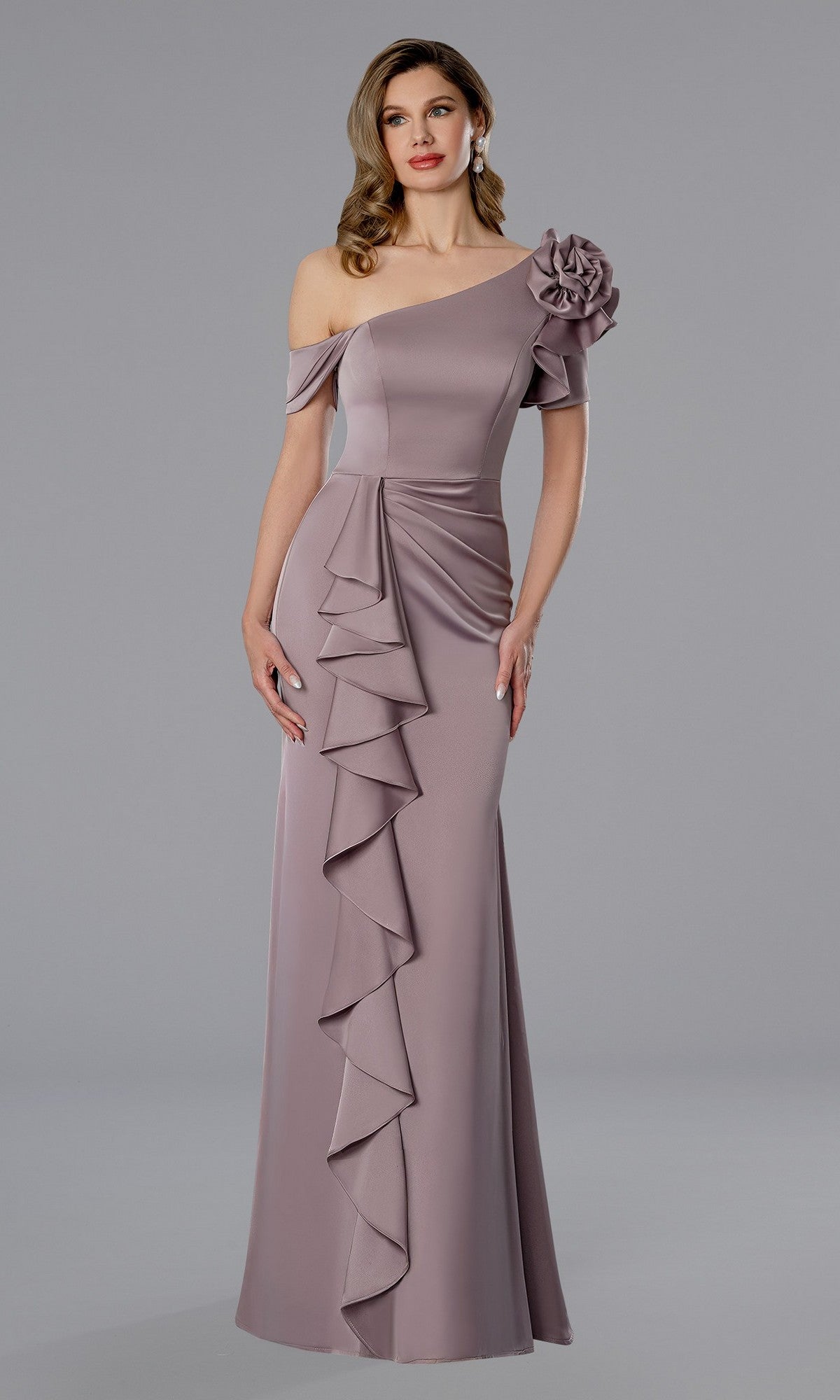 A woman wears the Stella Couture 25360 long formal dress in rose, featuring an asymmetrical neckline, a bold 3D flower on one shoulder, and a cascading front ruffle.