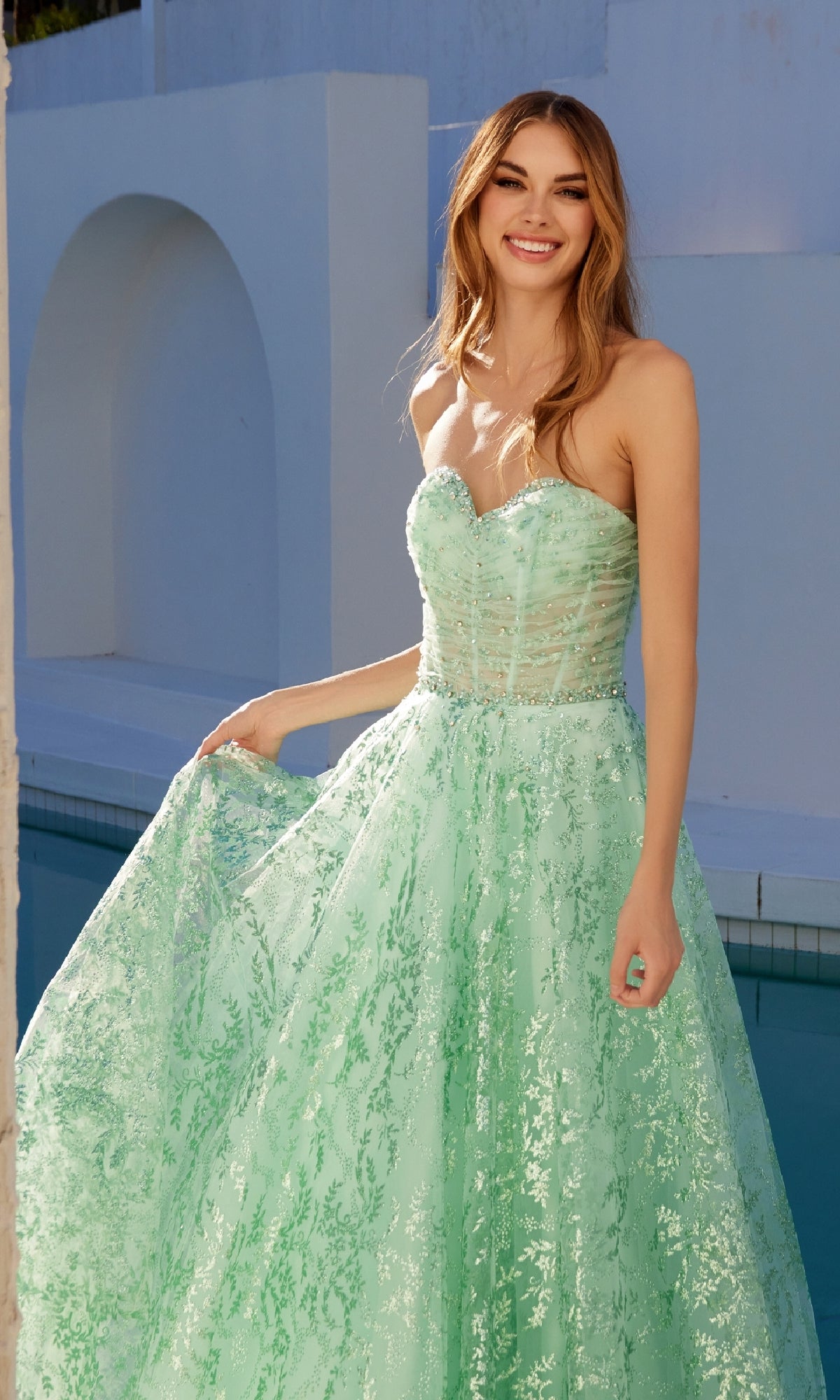 A woman stands by a pool, smiling in the Strapless Glitter-Print Prom Gown: Juliet JT2547S, featuring mint green shimmering patterned fabric.