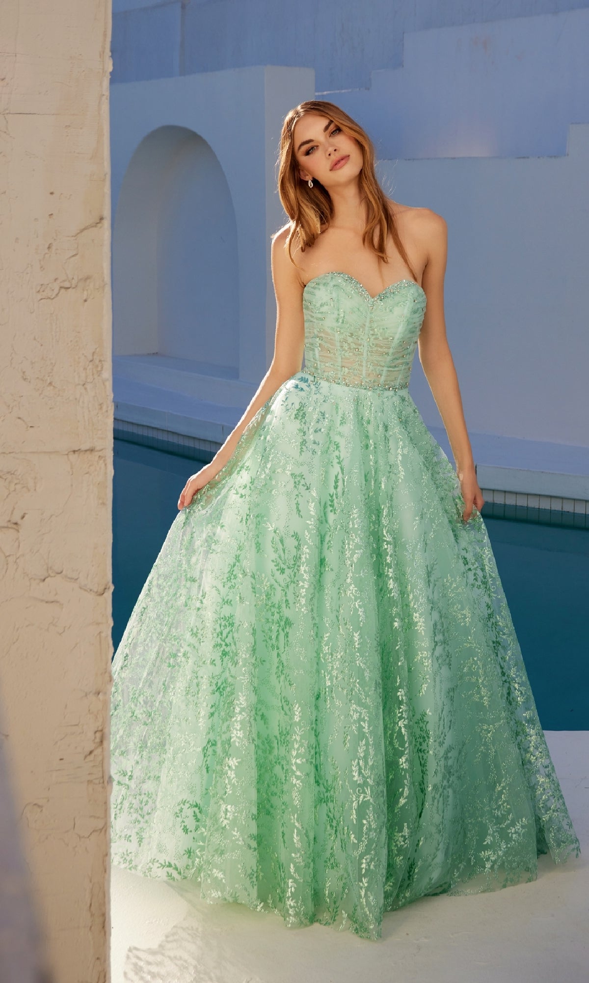 A woman stands outdoors by a pool, wearing the Strapless Glitter-Print Prom Gown: Juliet JT2547S—a mint green corset-style dress with a full skirt and lace details.