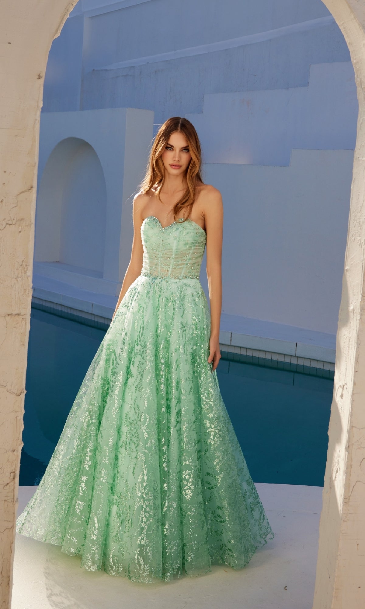 A woman stands by a pool wearing the Strapless Glitter-Print Prom Gown: Juliet JT2547S, a mint green dress with a fitted bodice and shimmering full textured skirt.