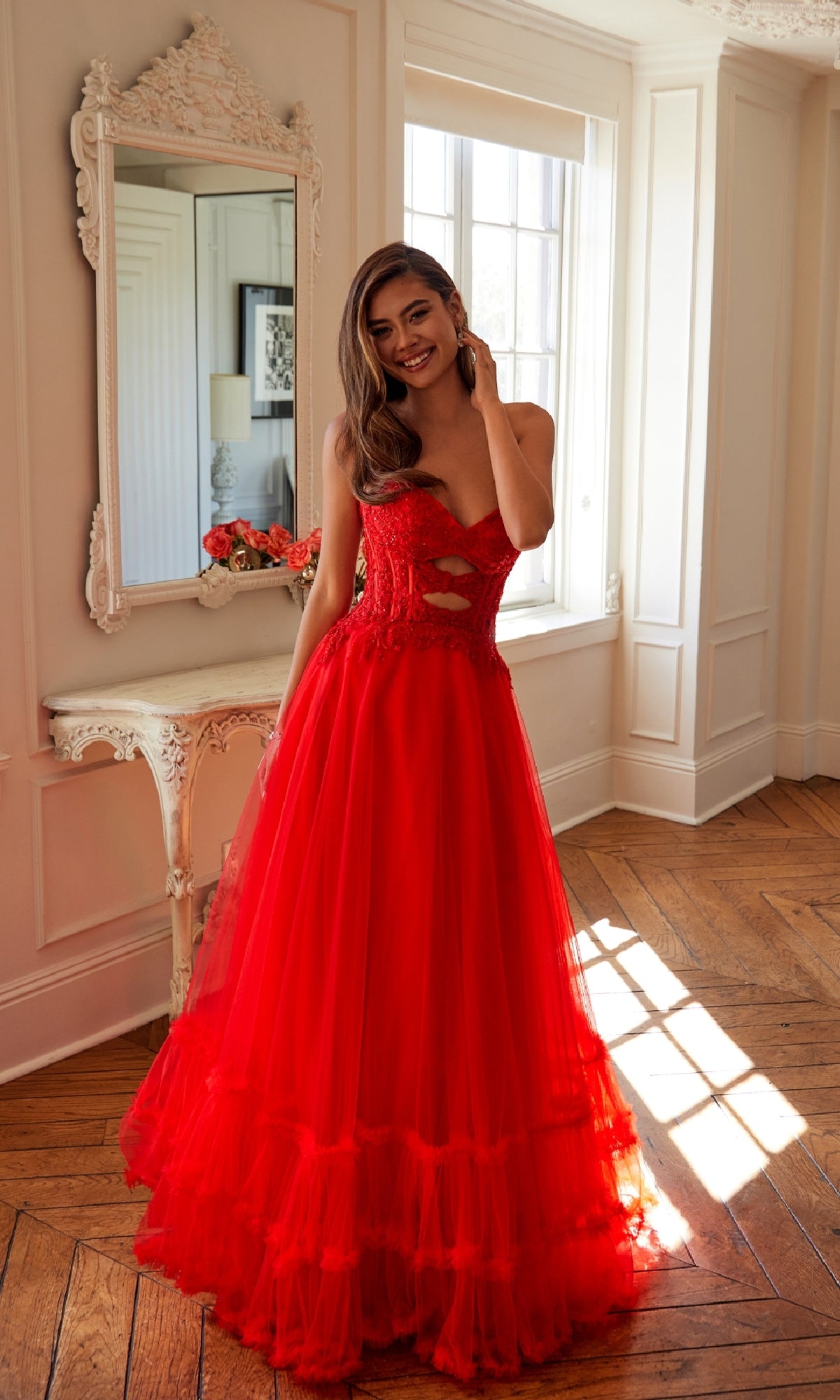 A woman in the Strapless Long Prom Ball Gown: Juliet JT2561S stands in a sunlit room with wooden floors, near a decorated mirror and a small table with flowers.