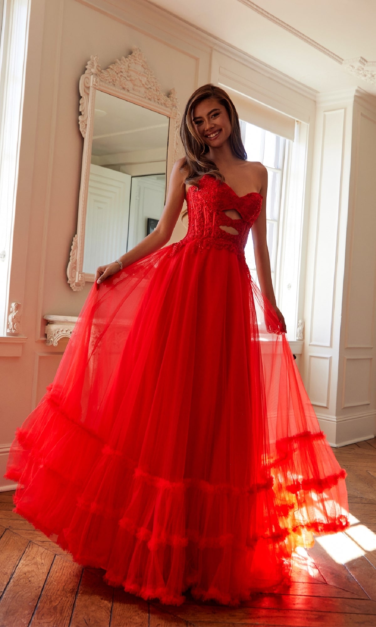 A woman in the Strapless Long Prom Ball Gown: Juliet JT2561S stands on a wooden floor, holding out the tulle skirt with one hand and smiling in a bright room with white walls and a decorative mirror.